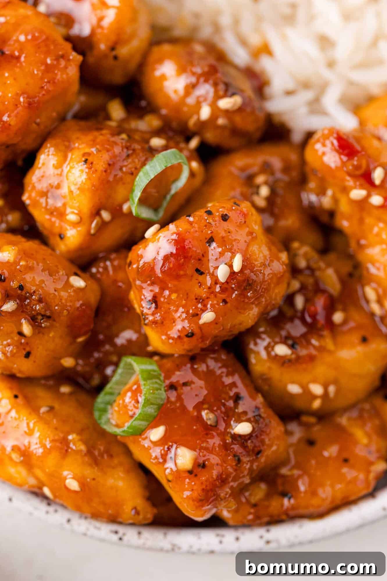 Close-up of saucy sweet chili chicken garnished with sesame seeds and green onions, served next to white rice.