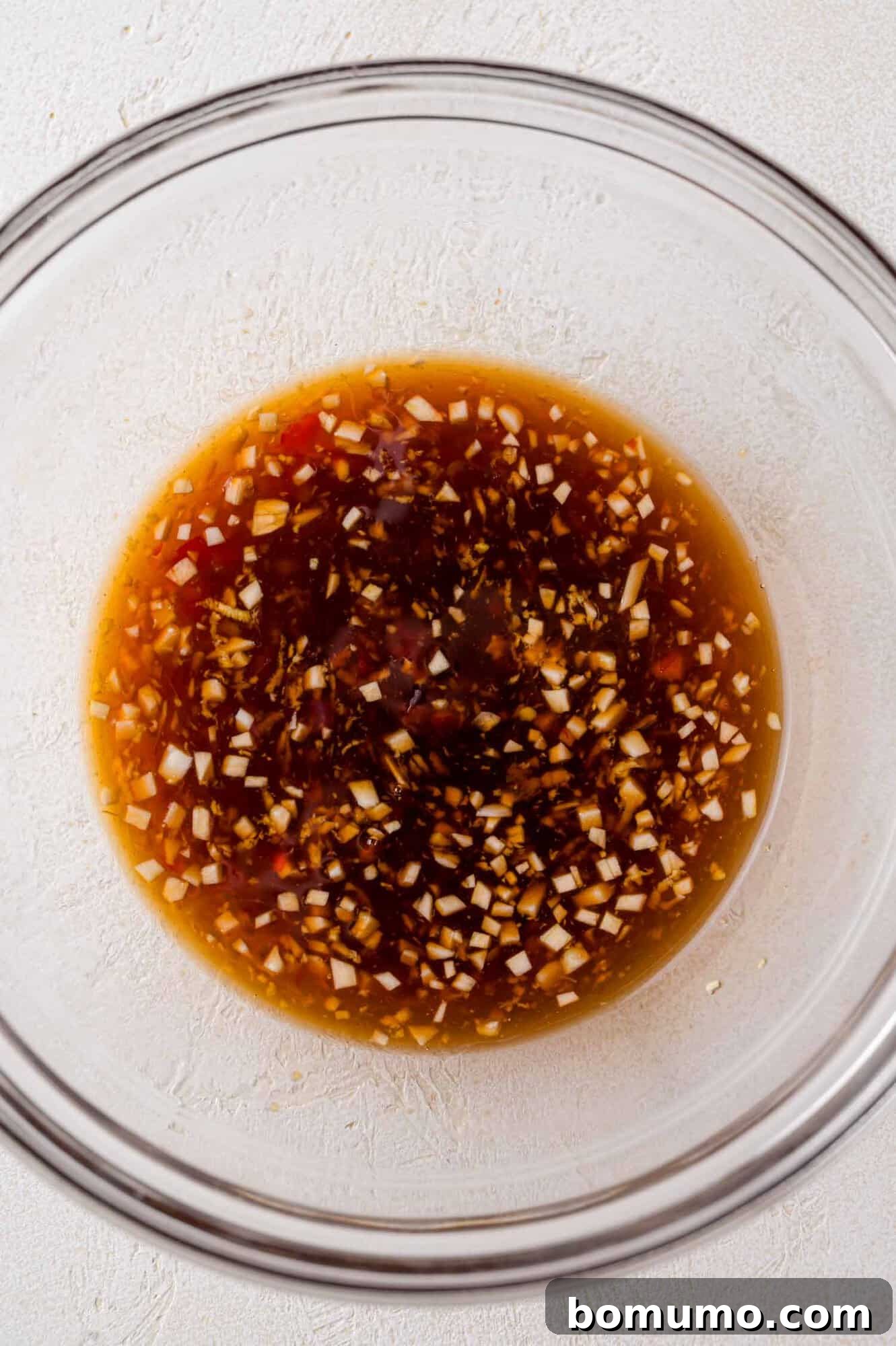 Close-up of a clear glass bowl containing a sauce mixed with finely chopped garlic on a white surface.