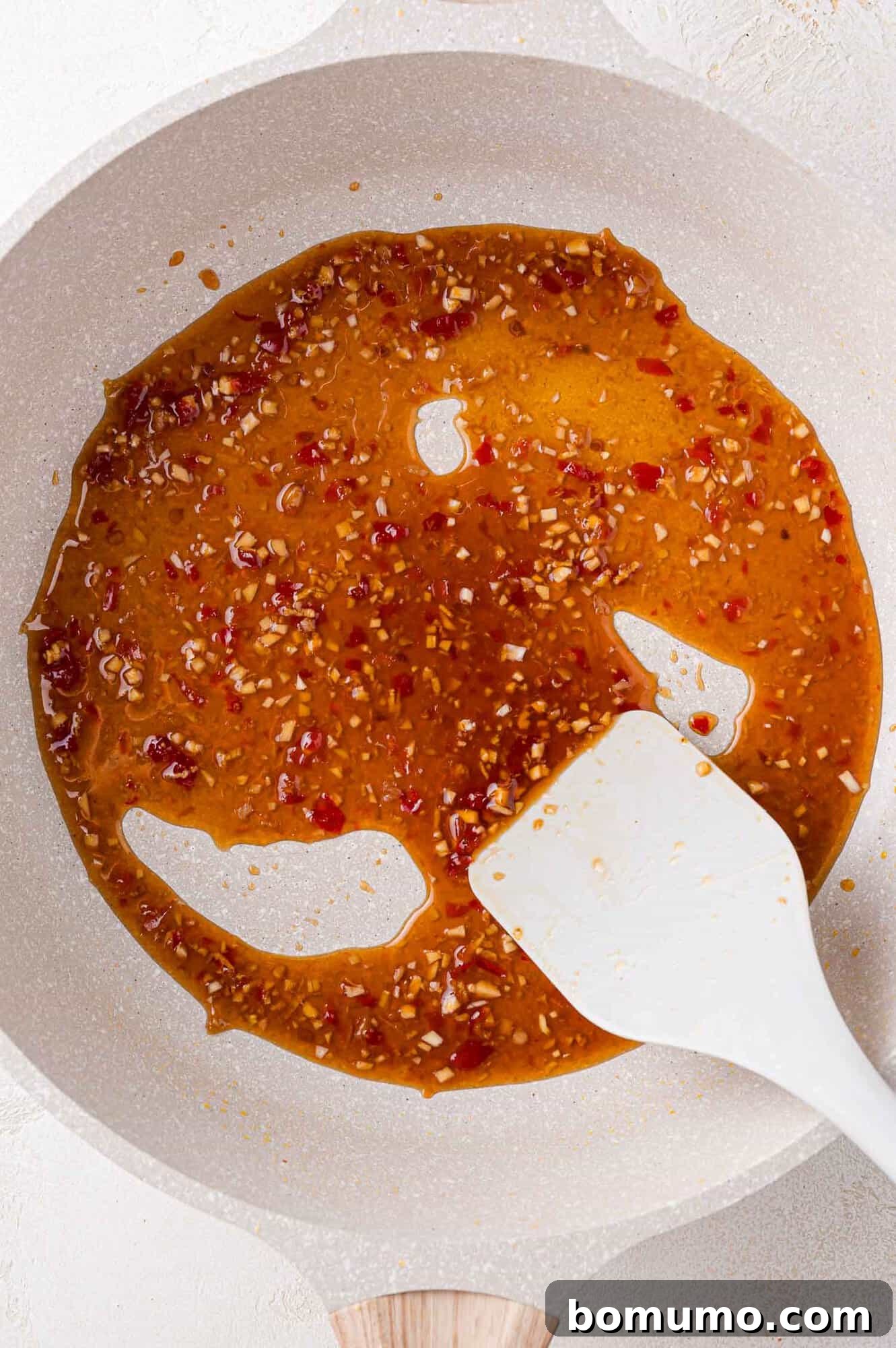 A white spatula stirring a mixture of brown sauce with red and white specks in a white pan.