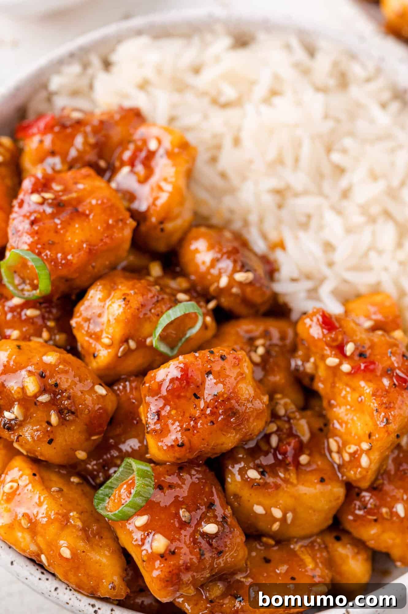 A bowl of white rice served with glazed chicken pieces garnished with sesame seeds and green onions.