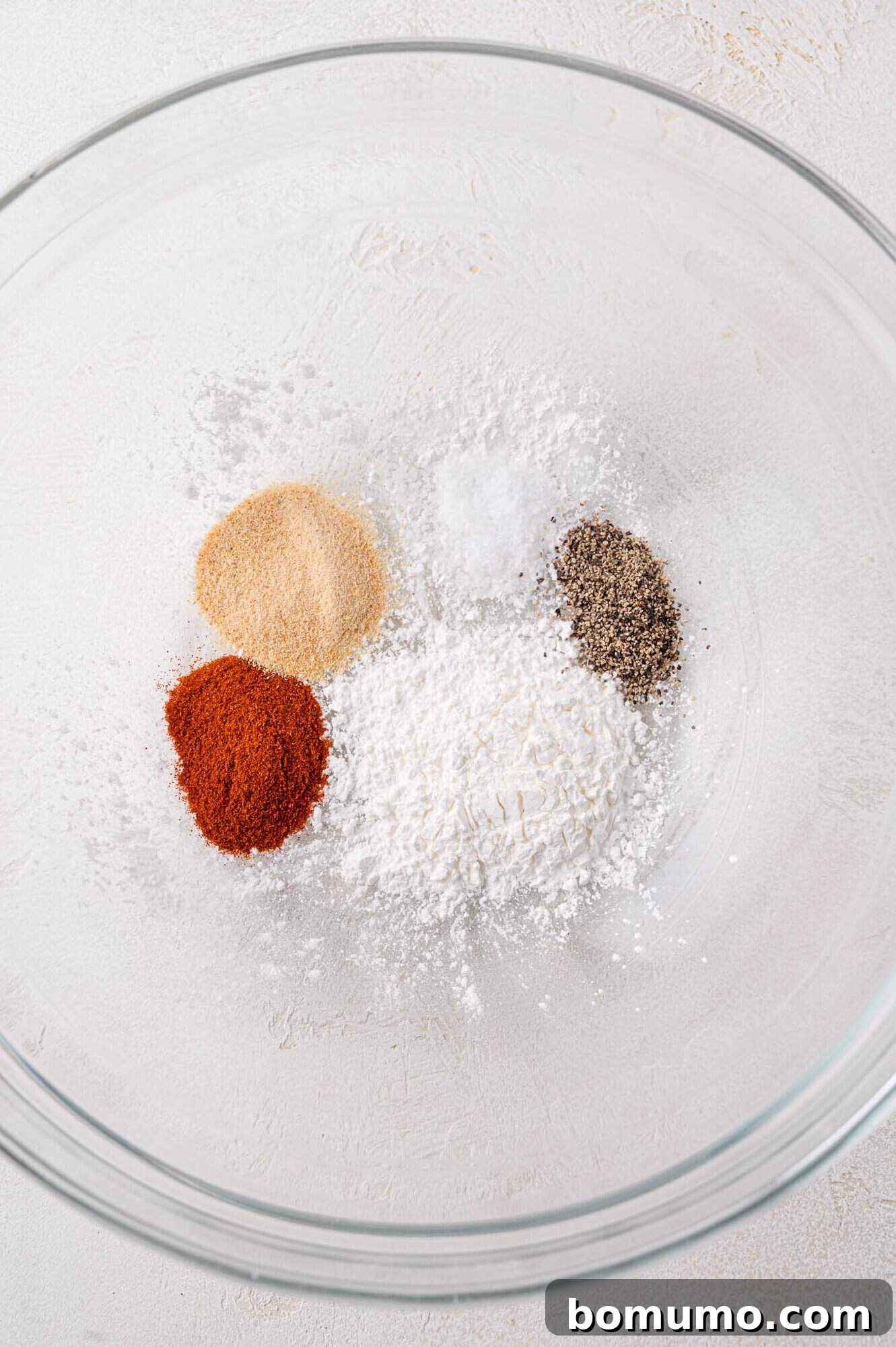 Glass bowl containing paprika, garlic powder, salt, pepper, and cornstarch on a textured white surface.