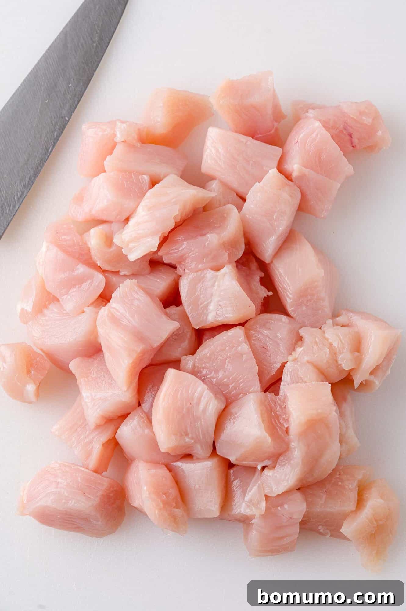 Diced raw chicken pieces on a white cutting board next to a chefs knife.