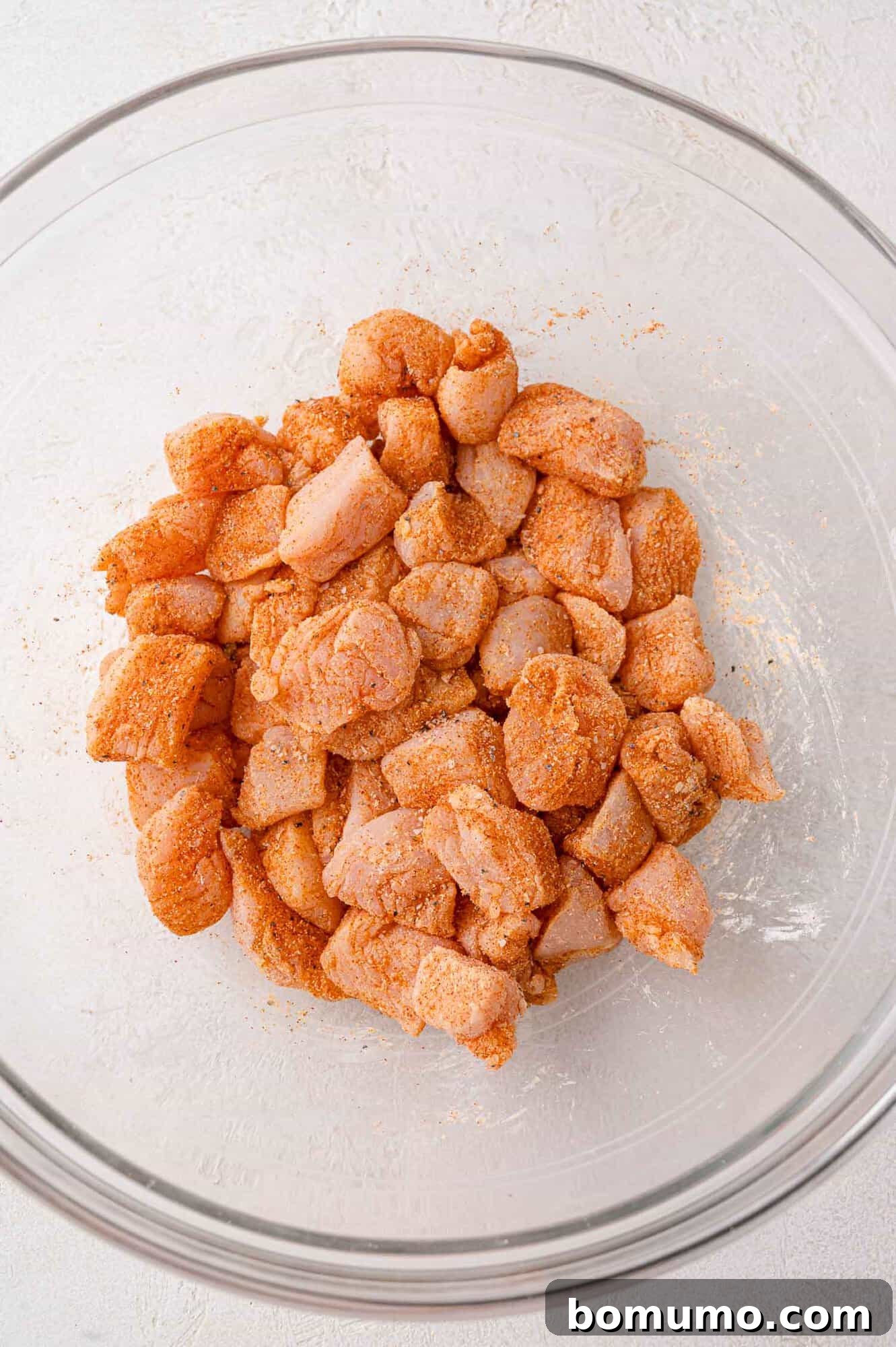 Diced chicken pieces coated with reddish-orange seasoning in a clear glass bowl.