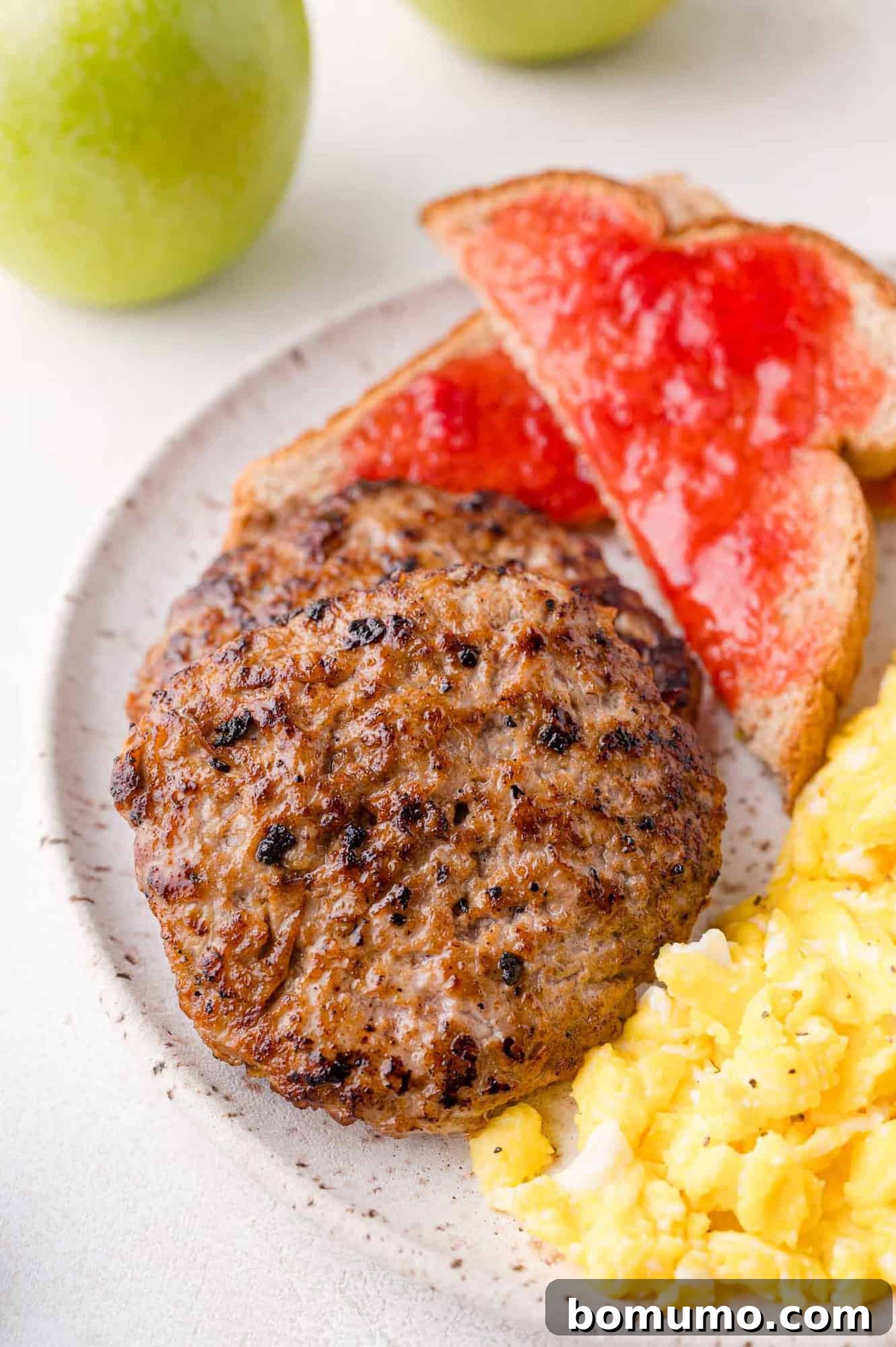 A plate with two sausage patties, scrambled eggs, and two slices of toast with red jelly. Green apples are visible in the background.