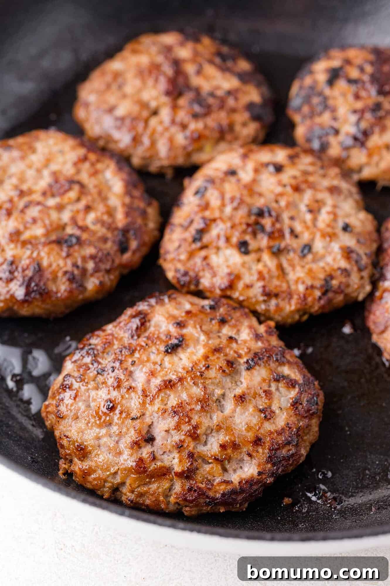 Six browned meat patties cooking in a black skillet, with visible browned marks and some oil in the pan.