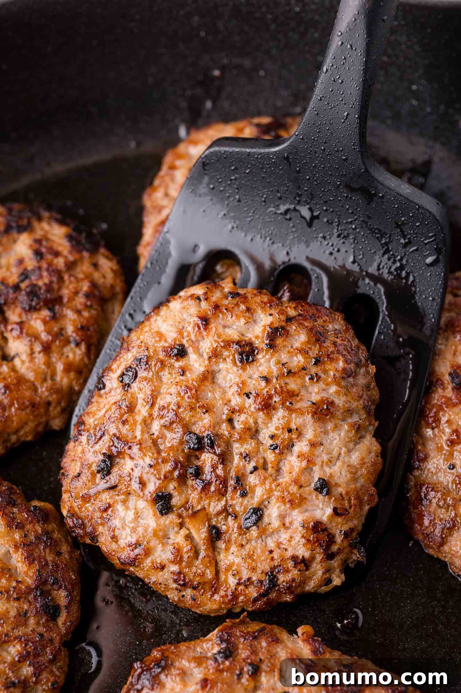 A close-up of a black spatula lifting a cooked, browned sausage patty from a skillet with several other patties.