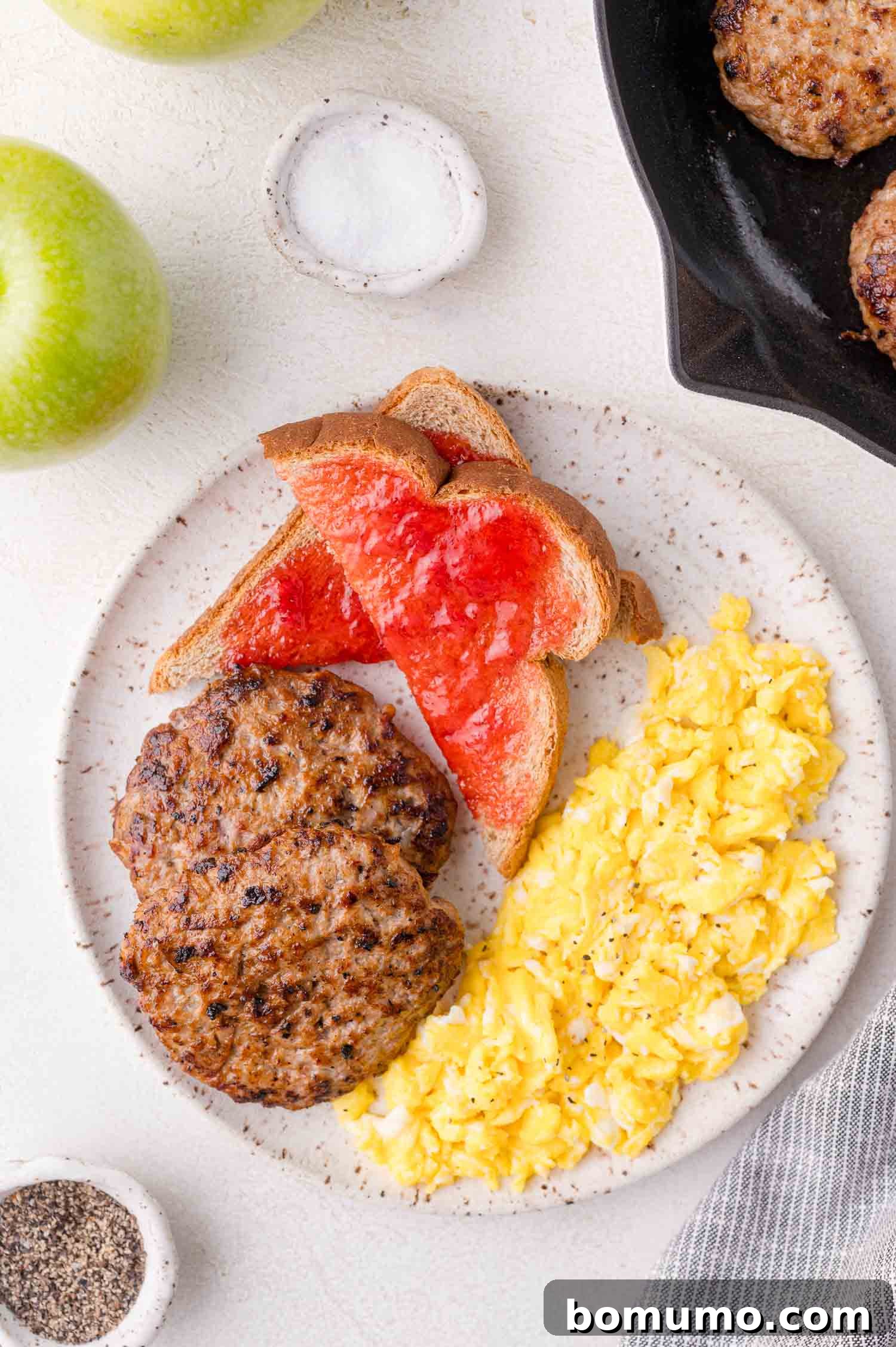 A plate with scrambled eggs, two sausage patties, and two slices of toast with red jelly. Green apples, salt, pepper, and a skillet are nearby.