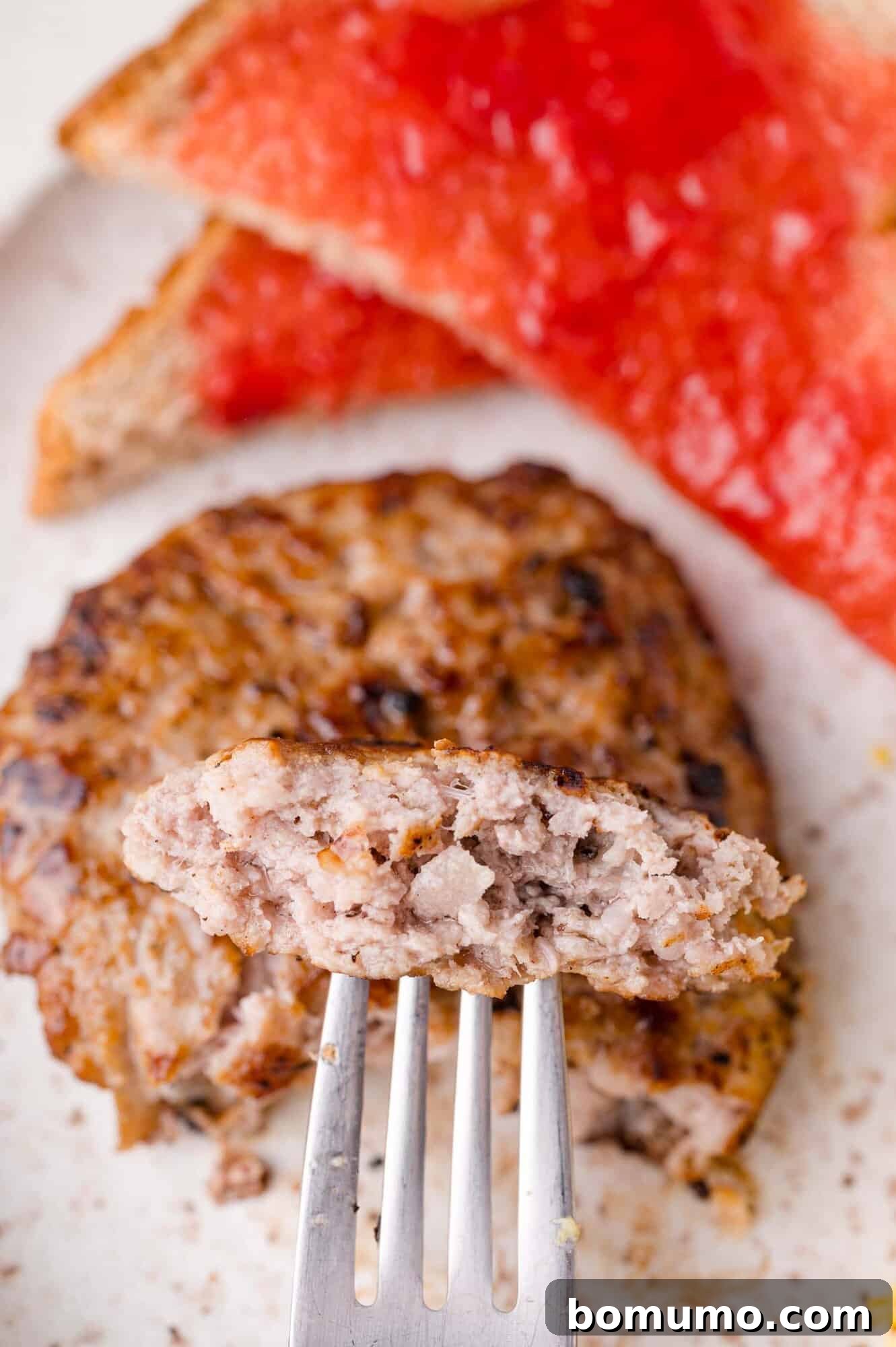 A close-up of a breakfast sausage patty with a piece on a fork, with toast topped with red jam in the background.