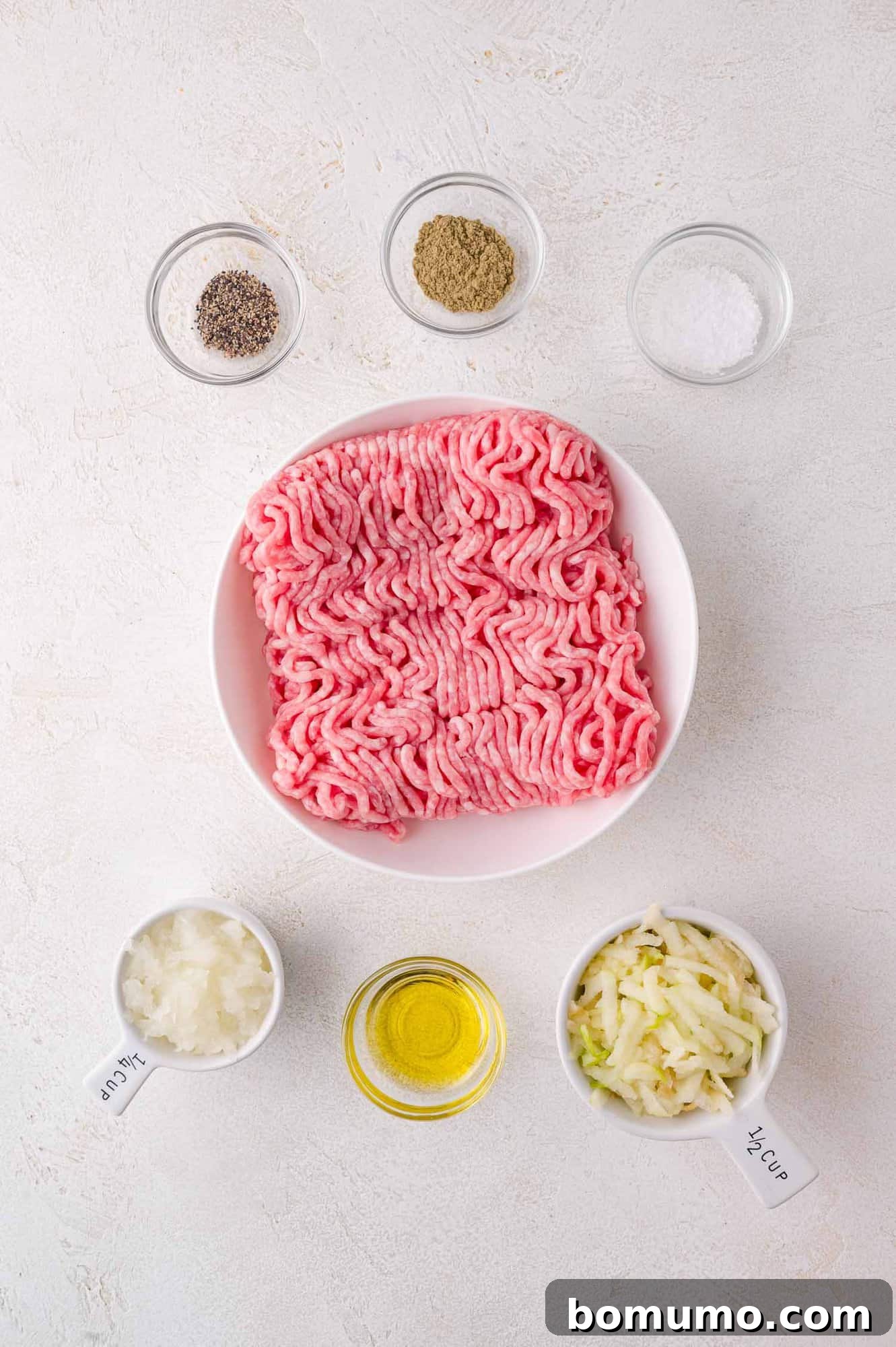 A bowl of raw ground pork, surrounded by small bowls of salt, pepper, spices, chopped onions, shredded apples, and oil on a white surface.