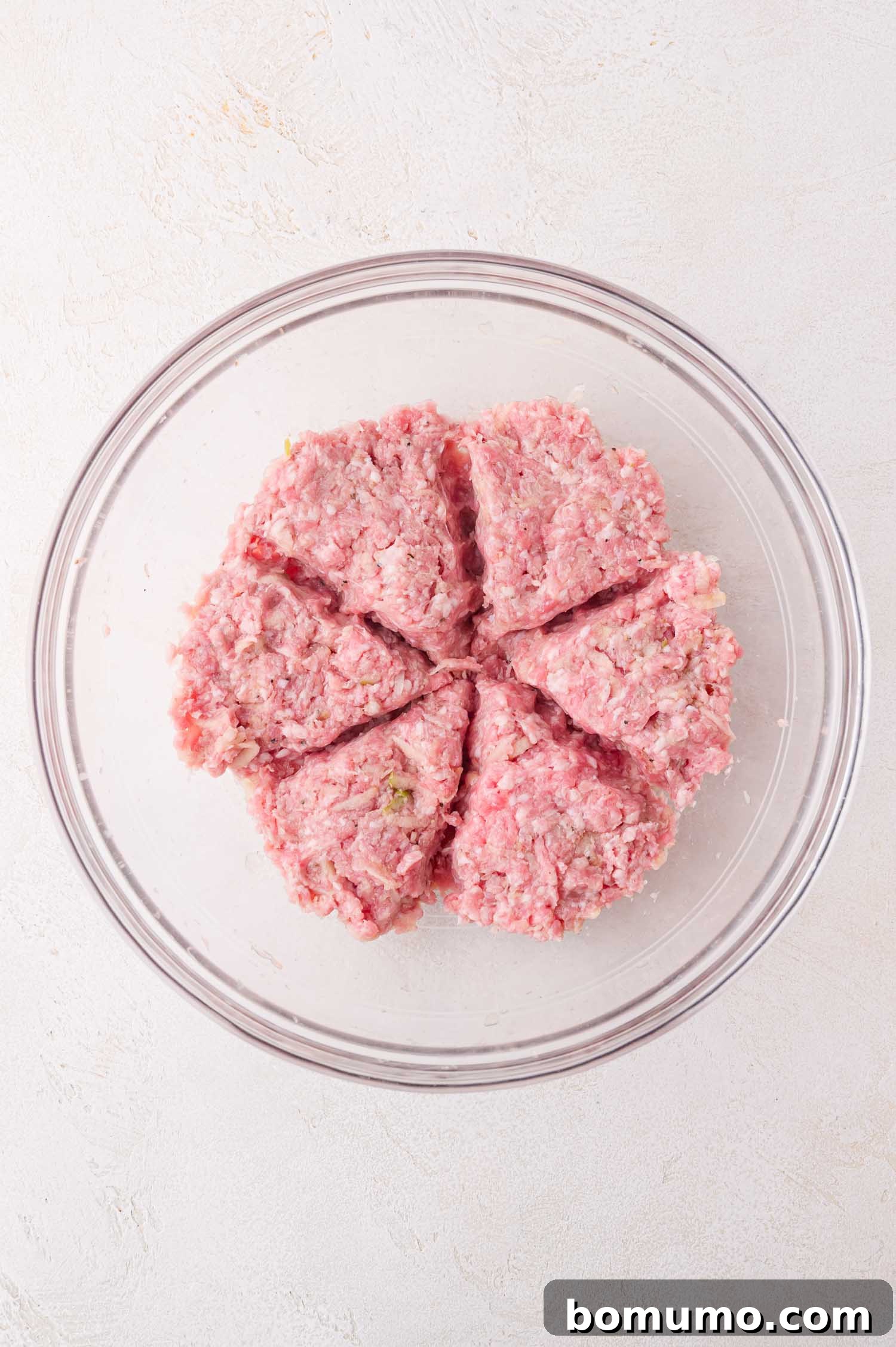 Raw ground meat divided into six equal portions in a clear glass bowl on a light-colored surface.