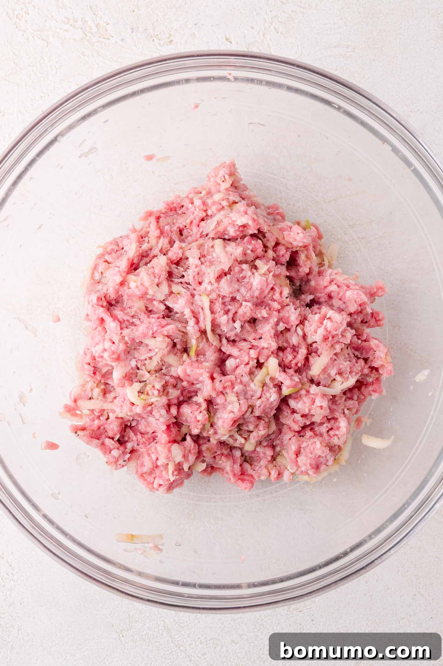 A clear glass bowl contains a mixture of raw ground meat and chopped onions on a light-colored surface.