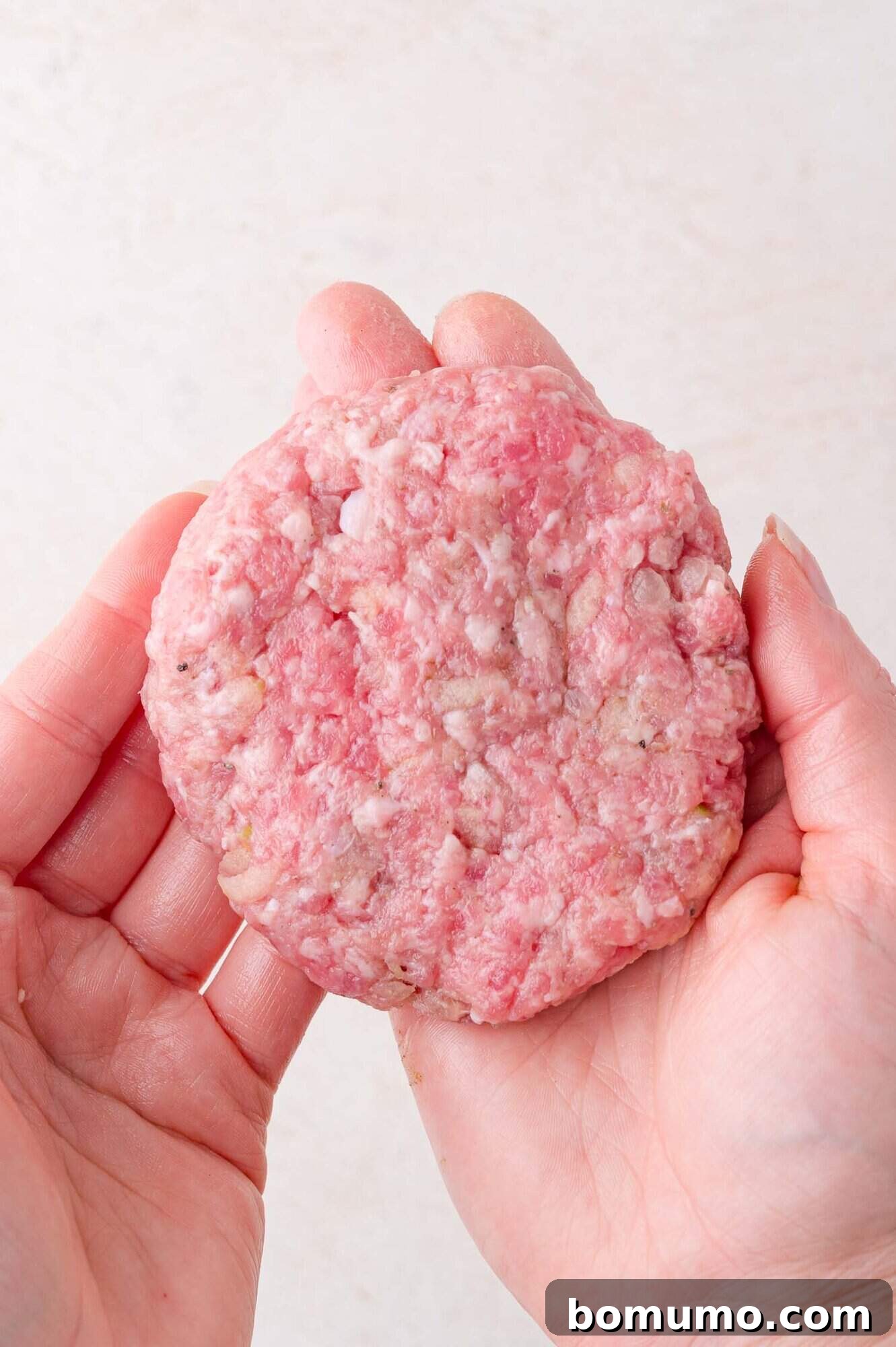 A pair of hands holds a uncooked sausage patty against a light background.