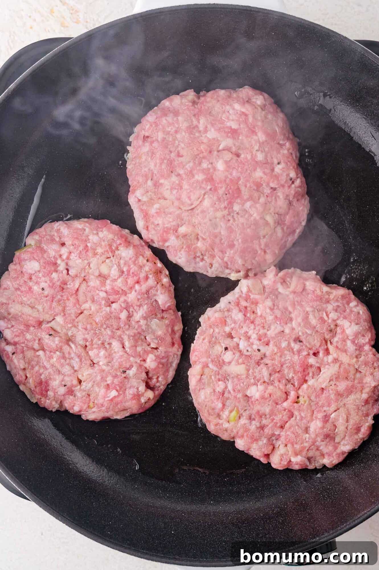 Three raw sausage patties are cooking in a black skillet with visible steam rising.