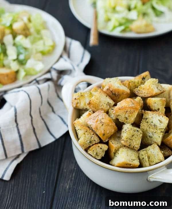 Effortless Homemade Caesar Croutons 3 A bowl filled with crisp Caesar croutons sits prominently, with two beautifully dressed Caesar salads blurred in the background, hinting at their delicious destination.