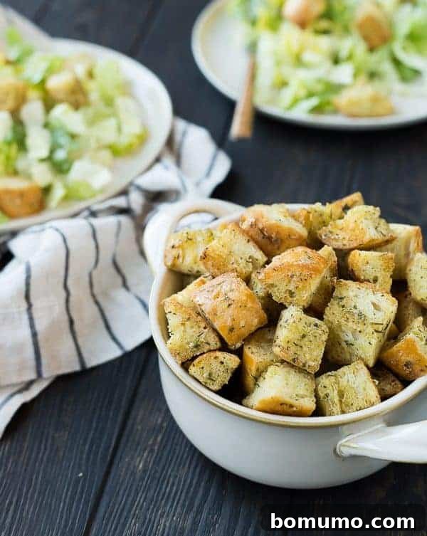 Effortless Homemade Caesar Croutons 5 A close-up view of golden Caesar croutons nestled in a white bowl, with tantalizing Caesar salads gently blurred in the background, highlighting the main attraction.