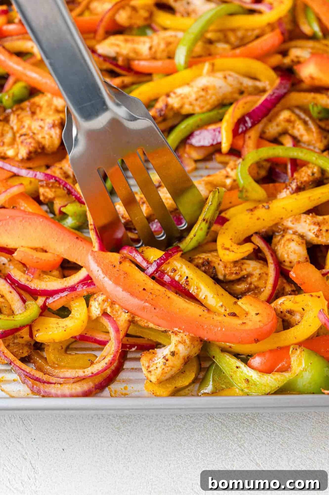 Tongs stirring cooked sheet pan fajitas on the pan to ensure even browning.