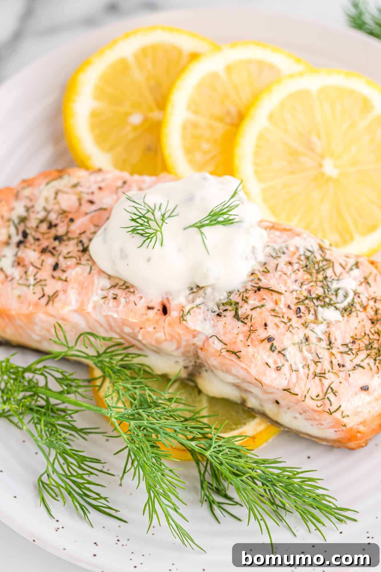 Lemon dill salmon topped with white sauce and fresh dill, next to lemon slices on a plate.