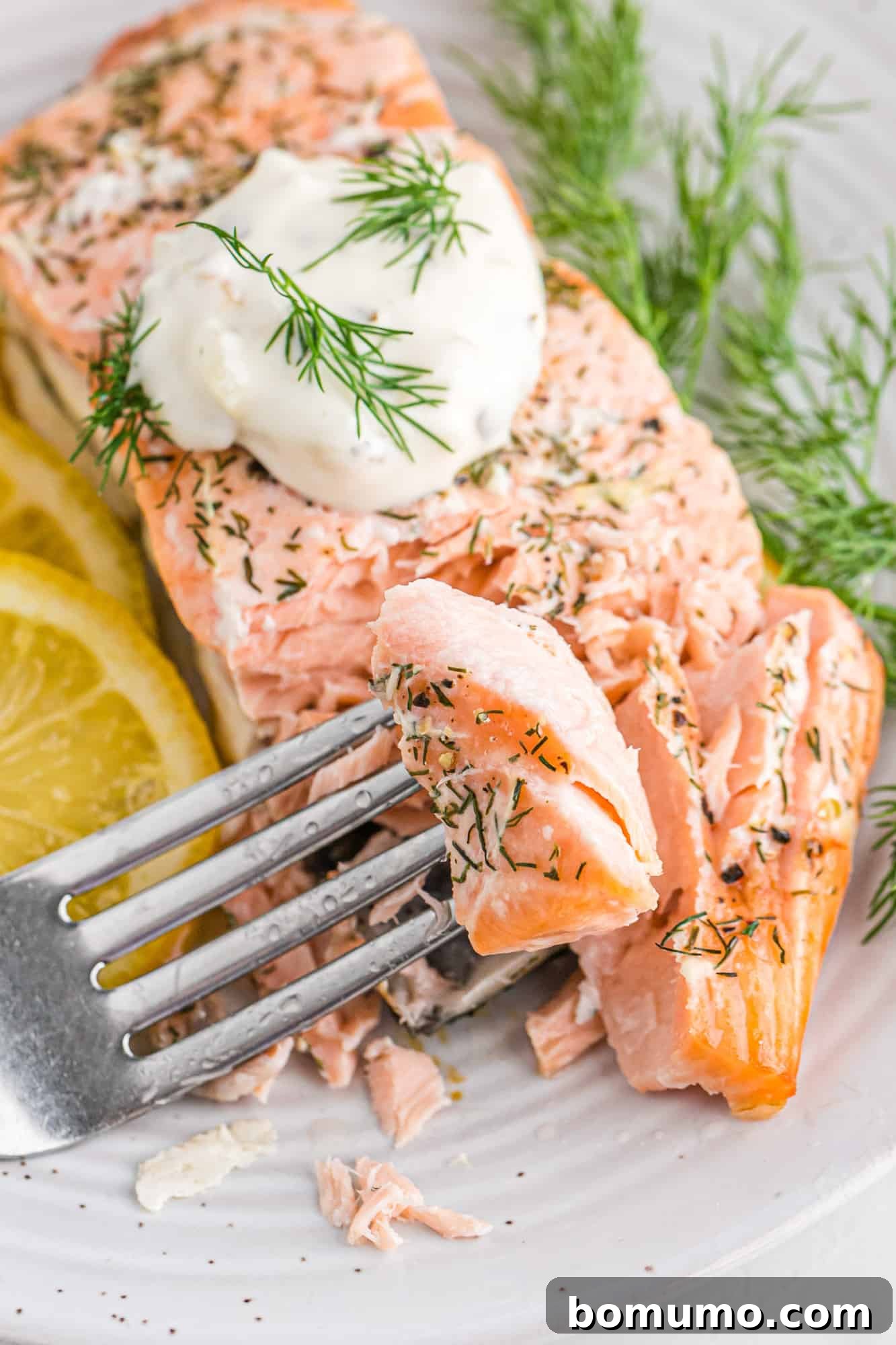 A fork lifting a piece of lemon salmon from a fillet topped with sour cream and dill sprigs.
