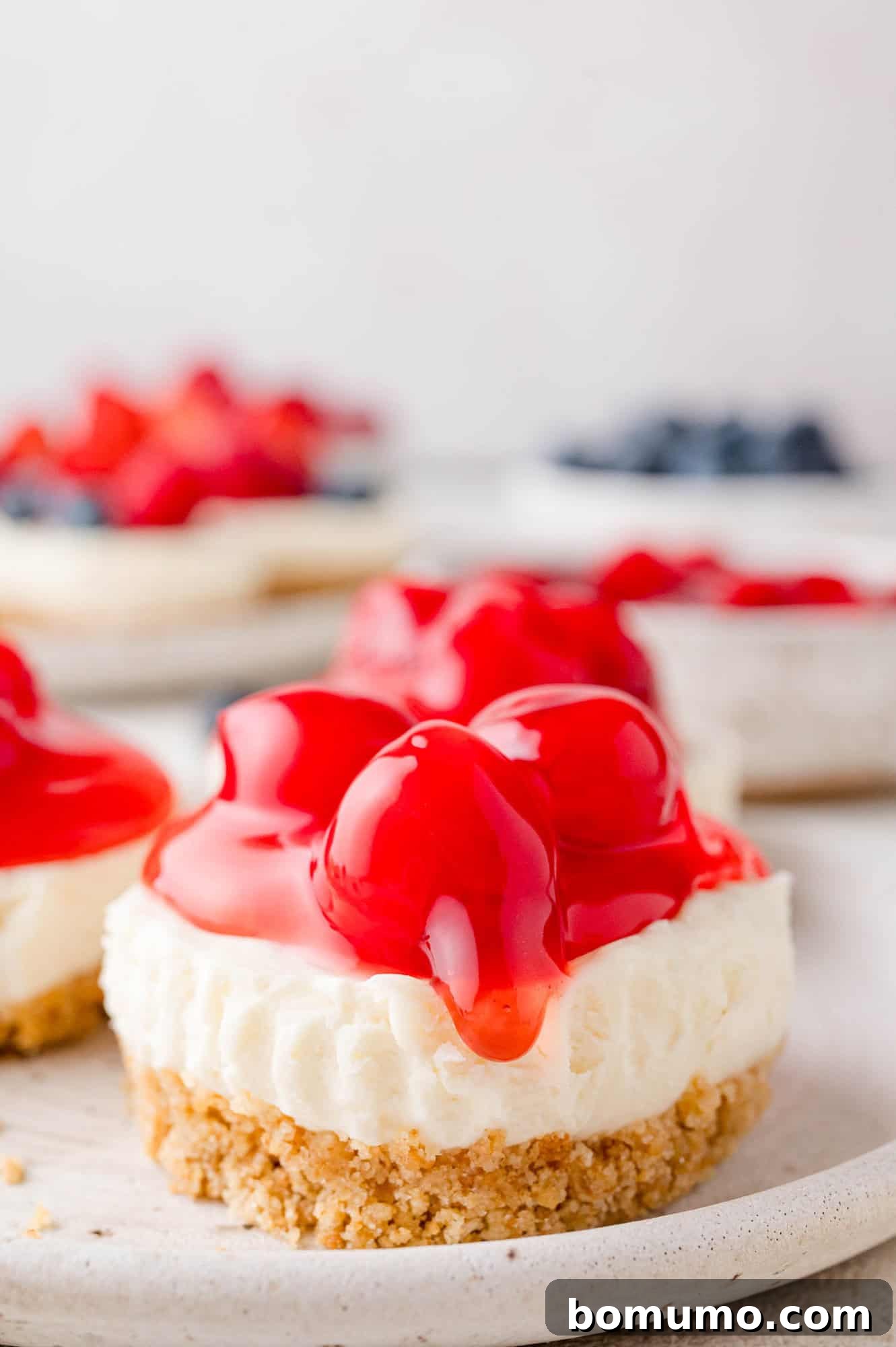 Effortless Mini Cheesecake Delights 2 Close-up of a mini cheesecake topped with glossy red cherry filling on a white plate.