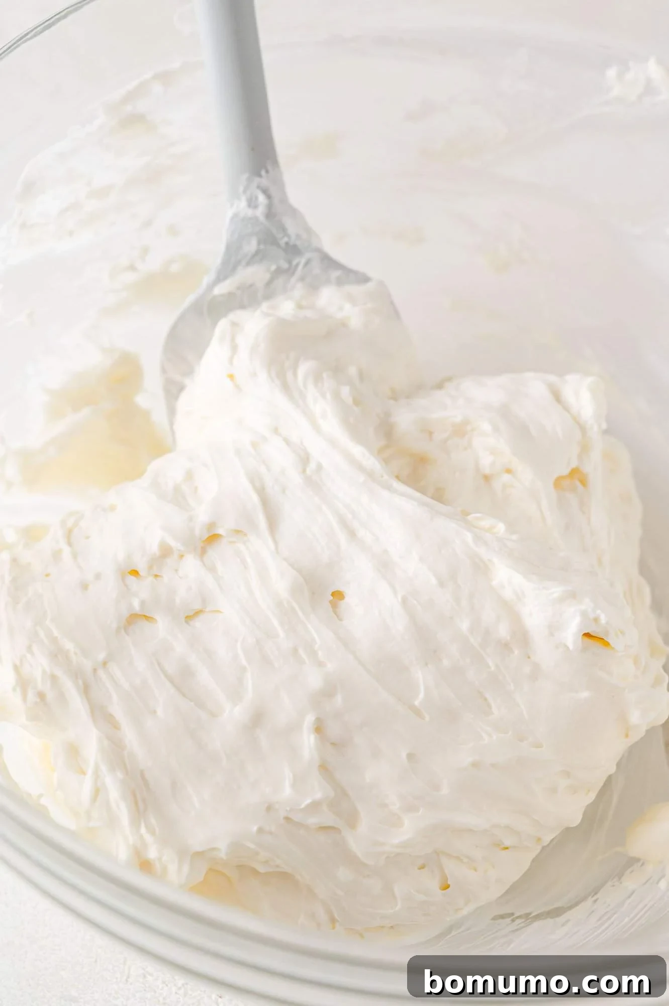 Effortless Mini Cheesecake Delights 13 A glass bowl containing thick, white whipped cream with a grey spatula.