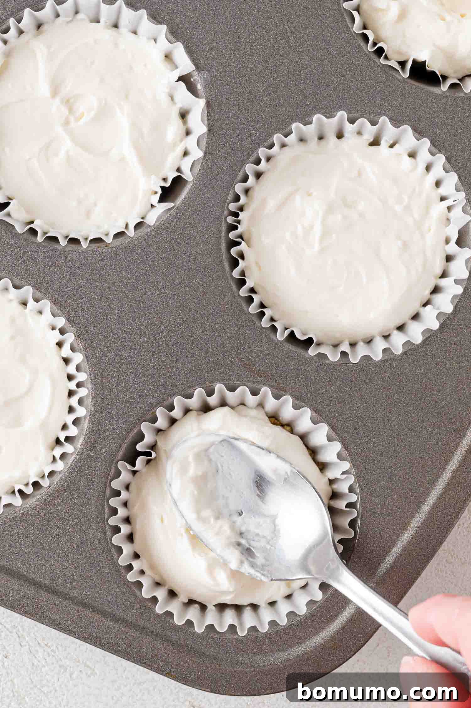 Effortless Mini Cheesecake Delights 14 Spoon spreading batter into cupcake liners in a muffin tin.