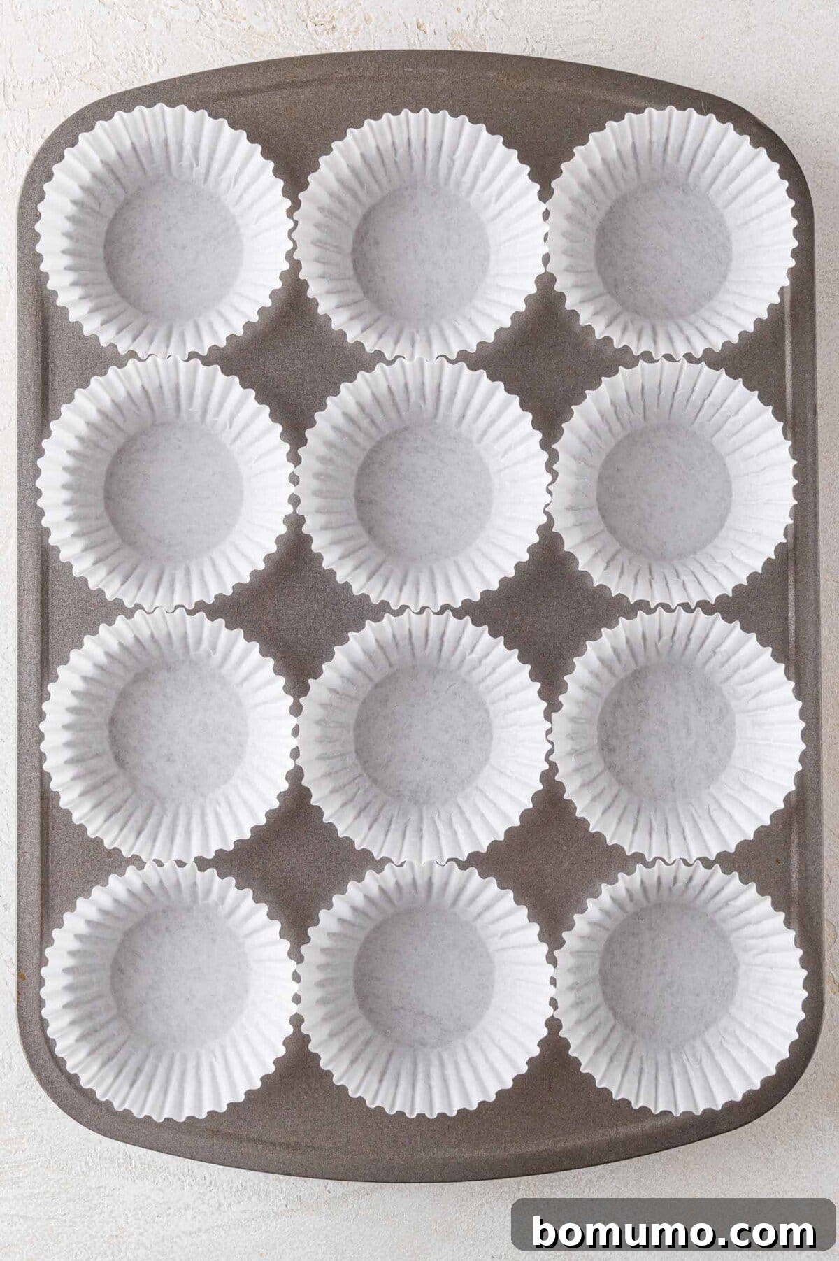 Effortless Mini Cheesecake Delights 8 A muffin tin with twelve white paper liners, arranged in a grid layout, on a light-colored surface.