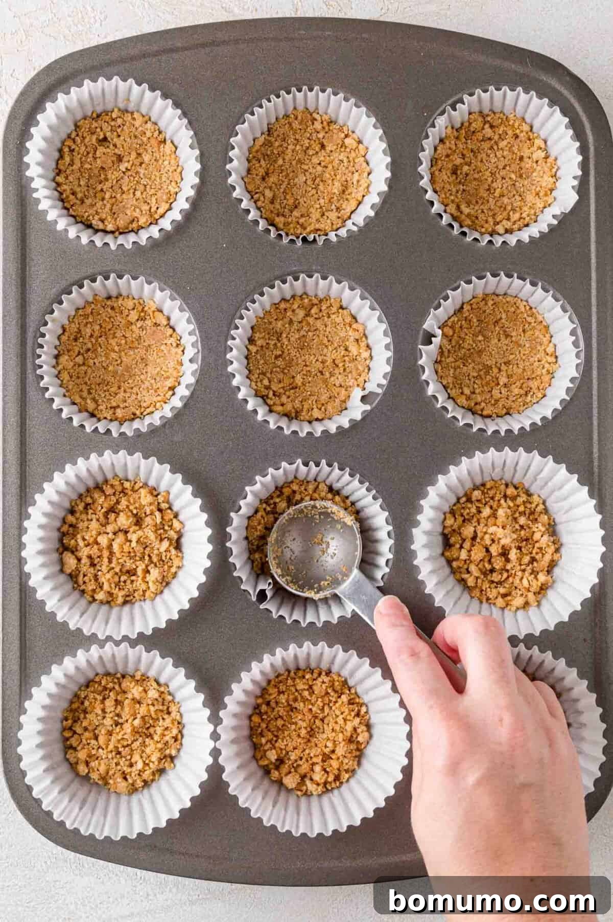Effortless Mini Cheesecake Delights 9 A hand presses graham cracker crumbs into cupcake liners in a muffin tin.