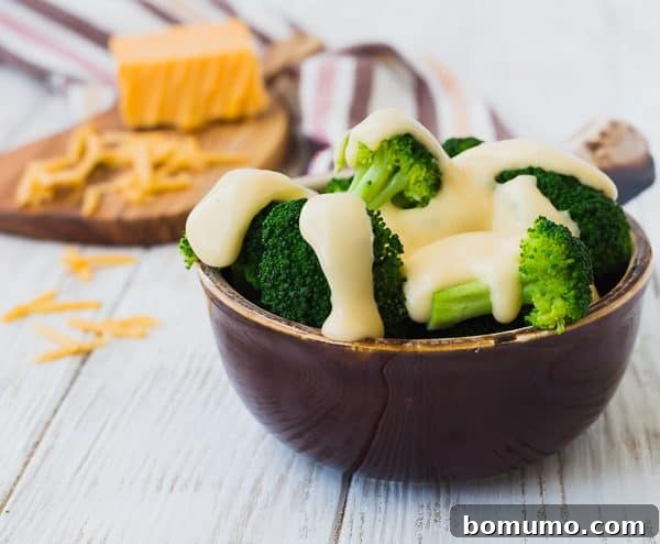 A bowl of steamed broccoli adorned with creamy cheese sauce, with a block of shredded cheddar cheese in the background.