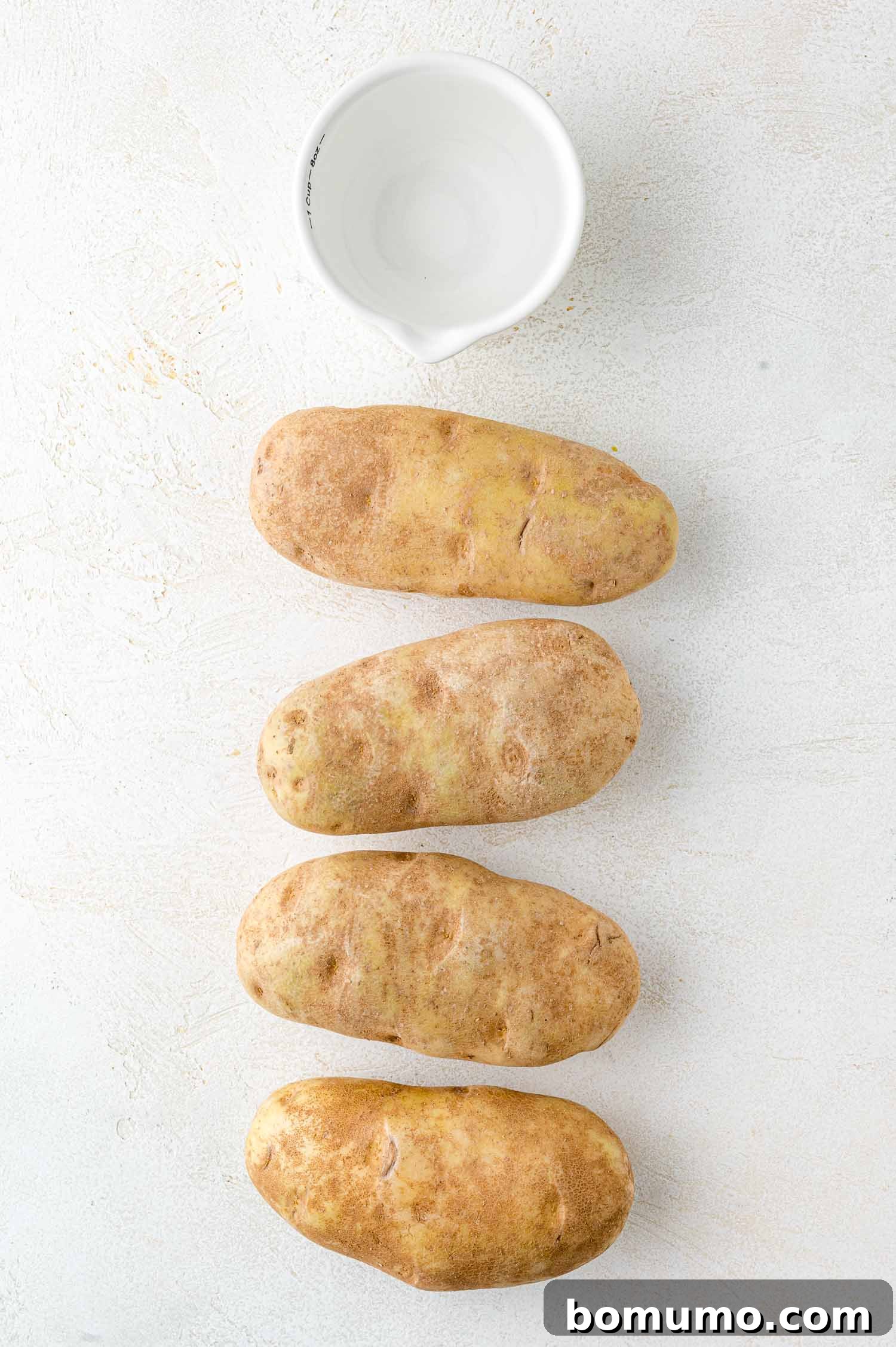 Foolproof Instant Pot Baked Potatoes 3 A selection of four clean russet potatoes arranged vertically on a light surface, ready for preparation. A small white bowl rests above them, hinting at the simple ingredients needed for this recipe.