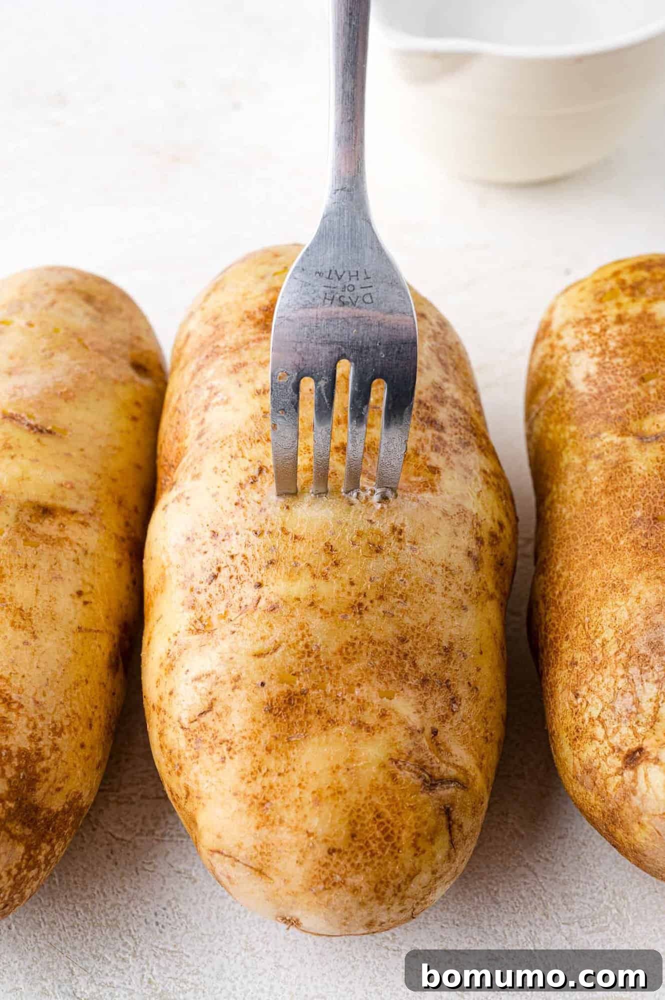 Foolproof Instant Pot Baked Potatoes 4 A metal fork delicately piercing the skin of one of three russet potatoes, laid out on a clean white surface. In the soft-focus background, a small white bowl suggests additional prep steps, illustrating the crucial pre-cooking piercing technique.