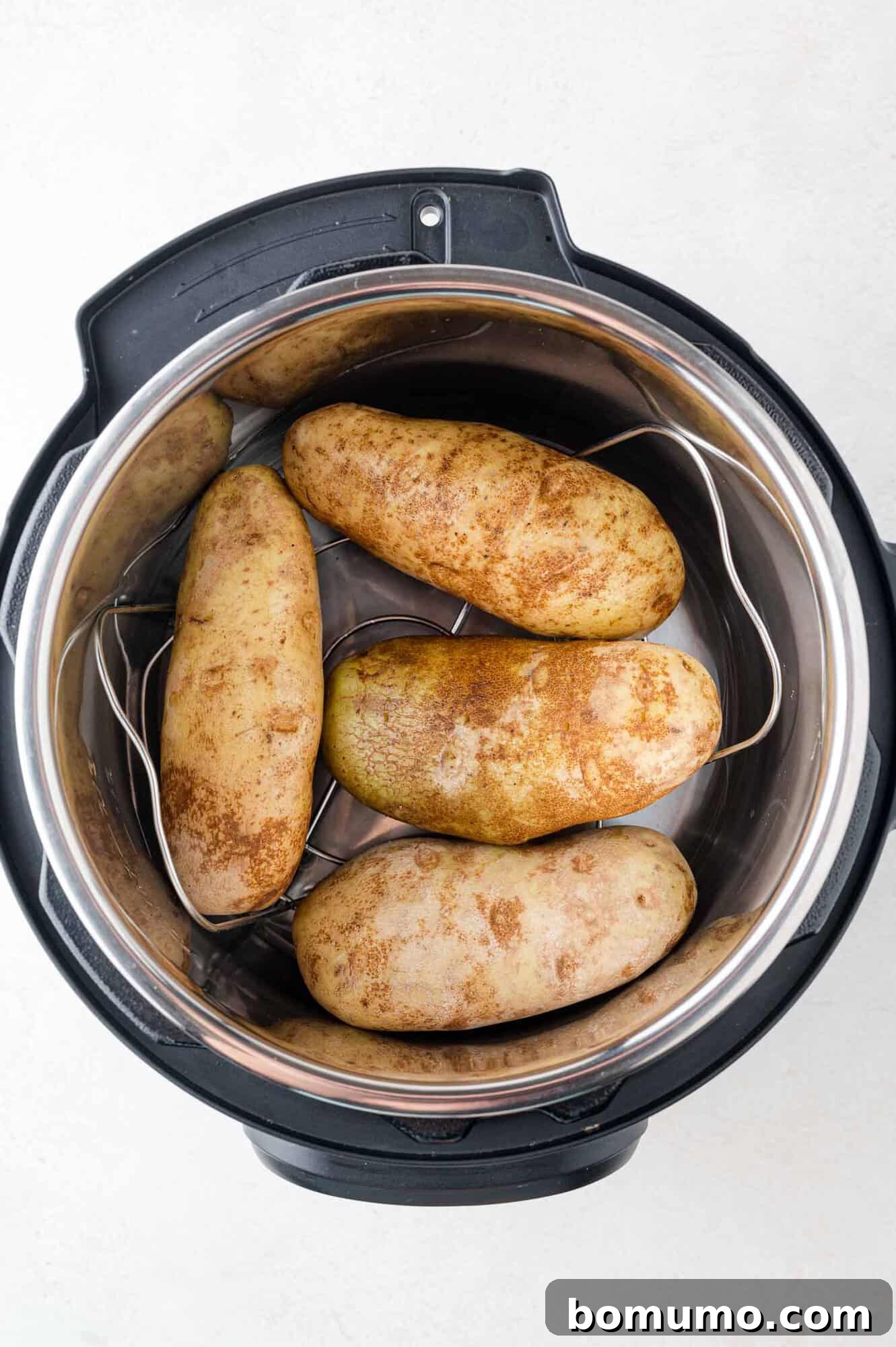 Foolproof Instant Pot Baked Potatoes 5 Four raw russet potatoes perfectly positioned on a metal trivet inside an Instant Pot, with a visible cup of water at the base, signifying readiness for pressure cooking. This image captures the precise setup for achieving evenly cooked results.