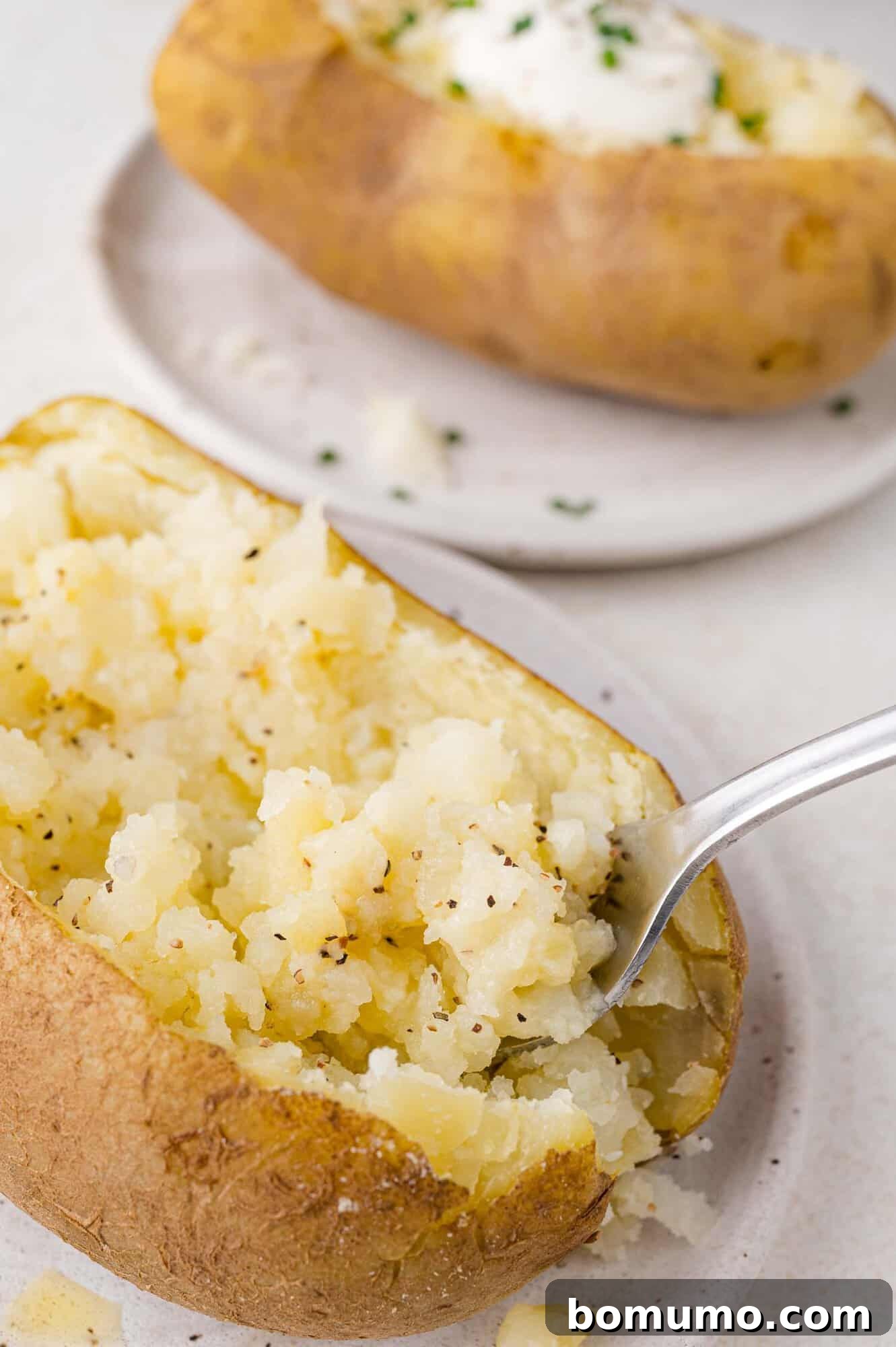 Foolproof Instant Pot Baked Potatoes 7 In the foreground, a perfectly baked potato is halved and its fluffy interior gently fluffed with a fork, ready for toppings. In the background, another potato is beautifully dressed with a dollop of sour cream and fresh herbs, artfully presented on a plate. This visual emphasizes both the ideal texture and serving potential of Instant Pot baked potatoes.