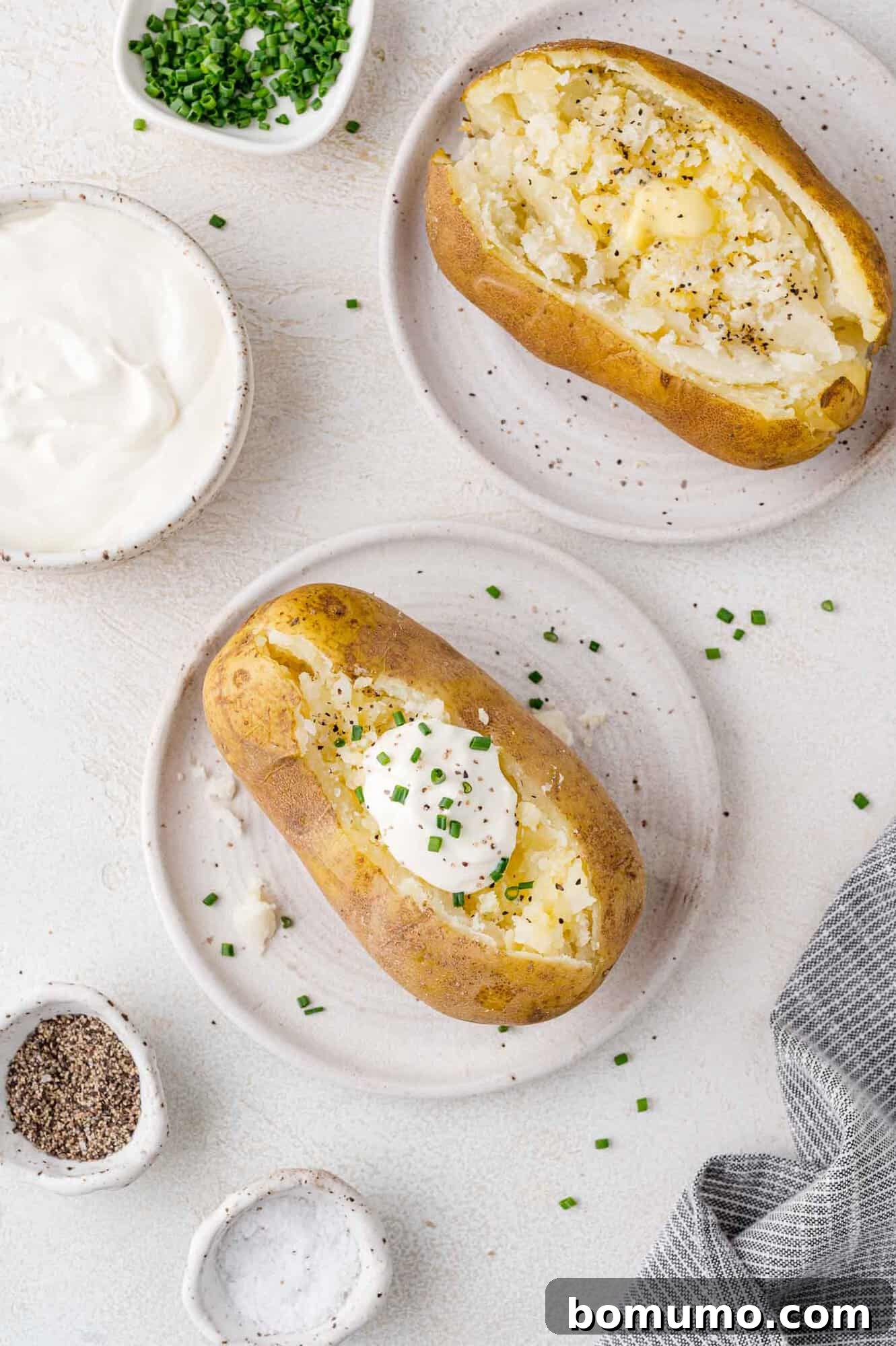 Foolproof Instant Pot Baked Potatoes 8 Two enticing baked potatoes presented on individual plates. One potato is lavishly topped with a dollop of sour cream and a garnish of fresh chives. Nearby, small white bowls hold additional sour cream, finely chopped chives, salt, and pepper, all arranged on a pristine white surface, ready for customization.