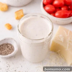 A jar of creamy Parmesan peppercorn dressing sits on a white surface, surrounded by grape tomatoes, a block of cheese, shredded cheese, black pepper, and croutons.