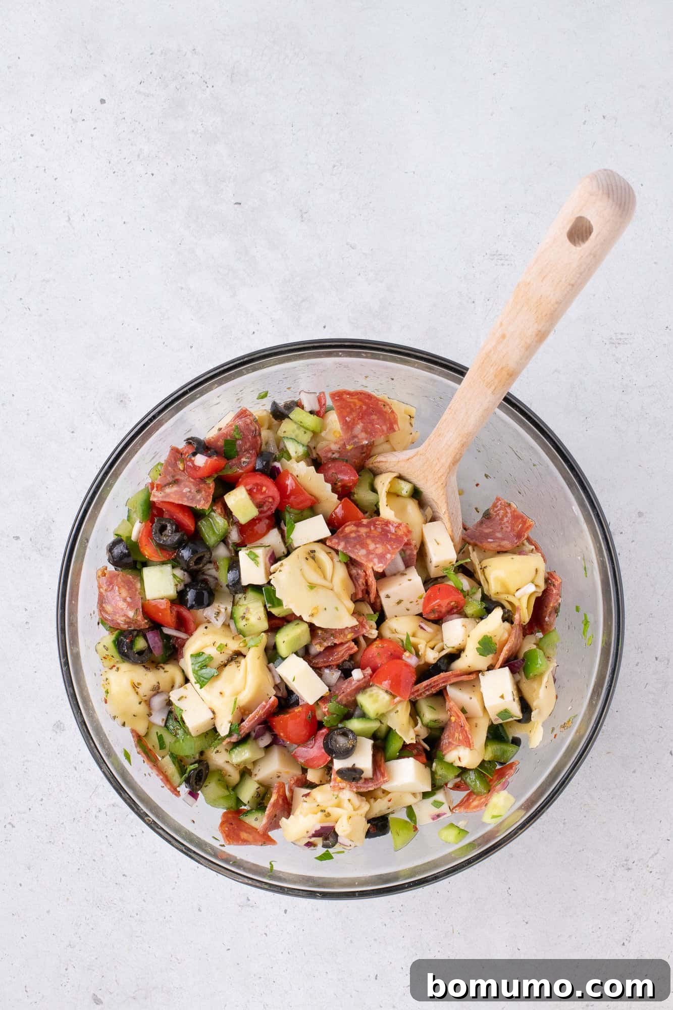 A glistening glass bowl filled with a fresh, chopped salad, featuring ripe tomatoes, crisp cucumbers, briny olives, savory cheese, and thinly sliced meat, accompanied by a rustic wooden spoon on a textured surface.