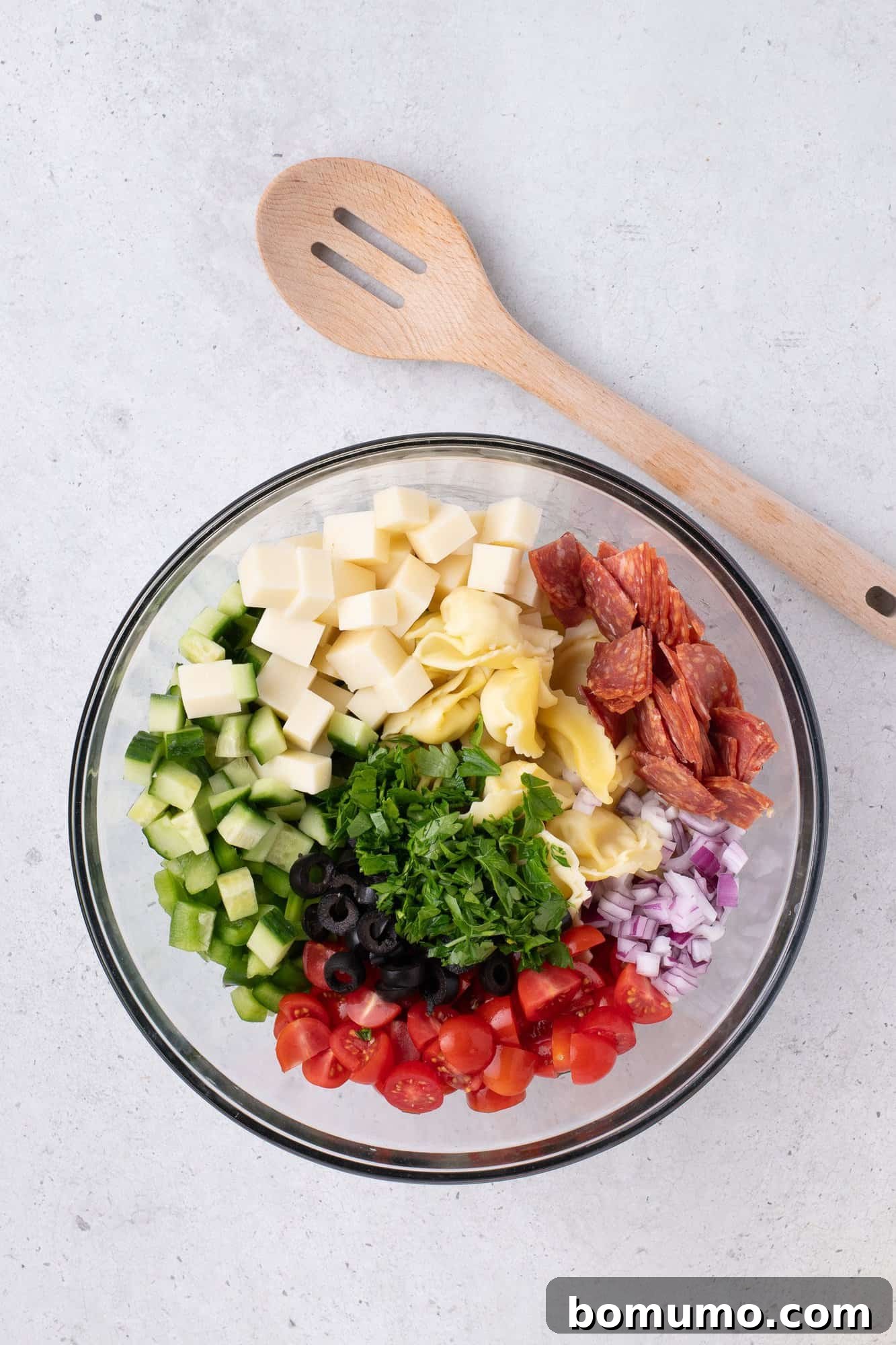 A large glass bowl overflowing with a colorful medley of chopped cucumbers, cubed mozzarella, cooked tortellini, pepperoni slices, black olives, halved grape tomatoes, diced red onion, and fresh parsley, with a wooden spoon resting nearby.