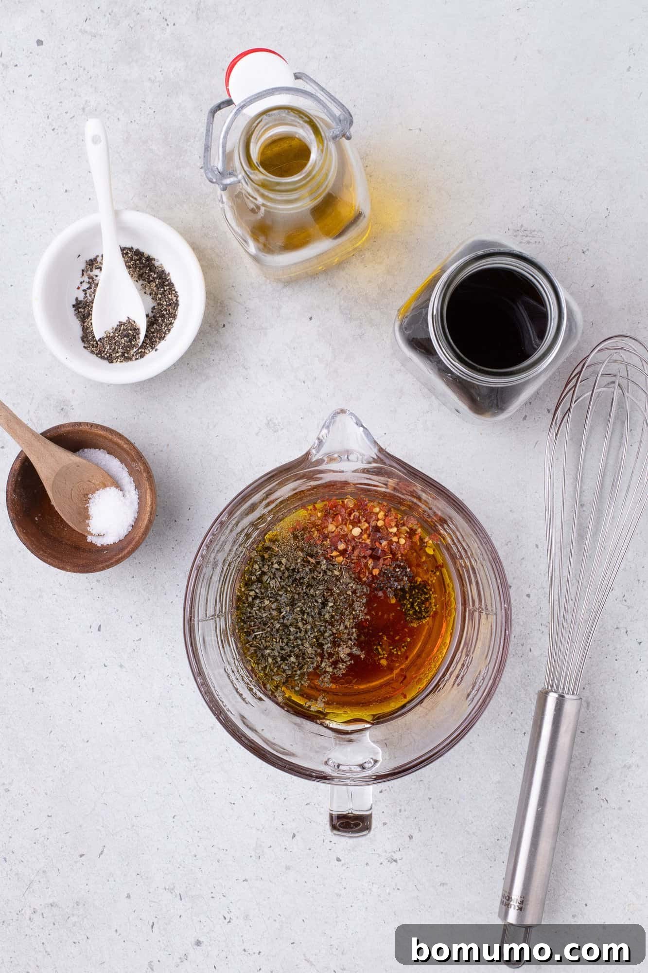 Dressing ingredients artfully arranged on a light surface, featuring a glass bowl of mixed oil and spices, a whisk, and individual small bowls containing salt, pepper, and fresh herbs. Two bottles of olive oil and red wine vinegar stand nearby.