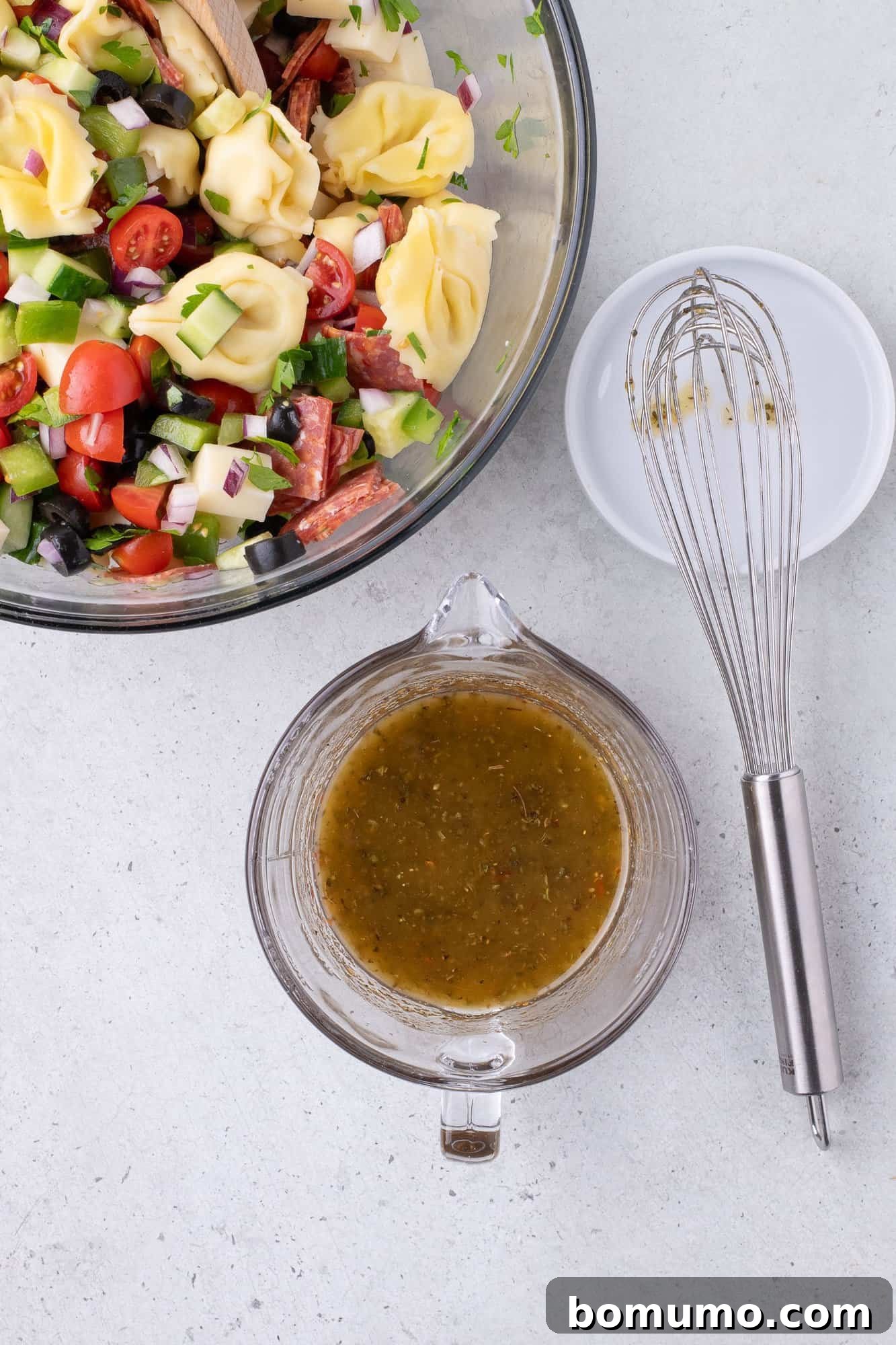 A vibrant bowl of tortellini pasta salad, brimming with cherry tomatoes, crisp cucumbers, colorful bell peppers, and briny olives, positioned alongside a whisk and a small bowl of freshly prepared dressing, ready to be combined.