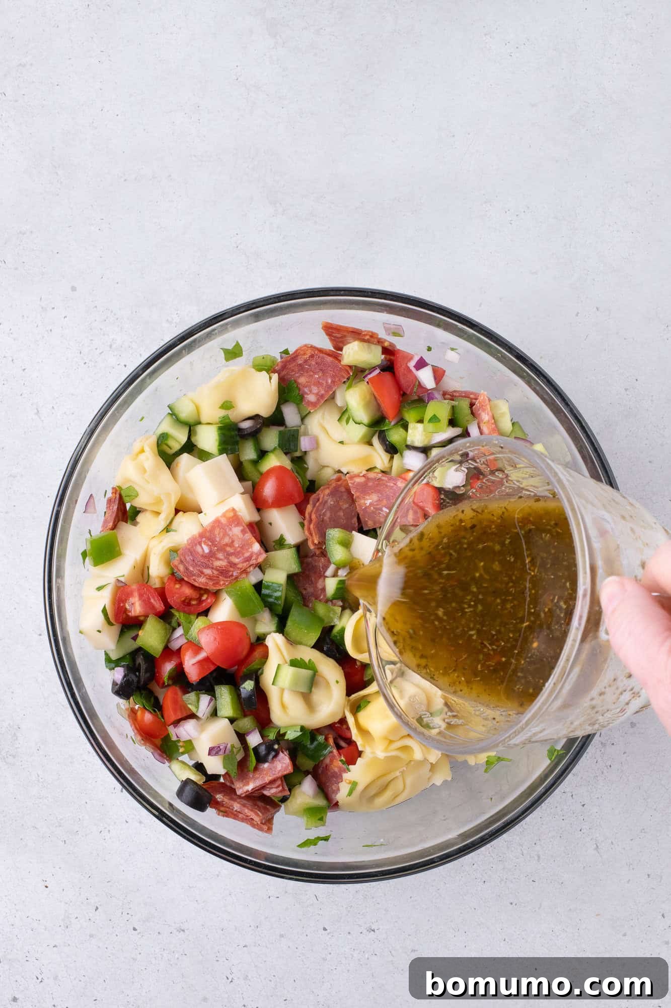 A hand elegantly pours a zesty dressing over a colorful tortellini salad, rich with salami, ripe tomatoes, creamy cheese, crisp cucumbers, black olives, and thinly sliced red onions, all nestled in a transparent glass bowl.