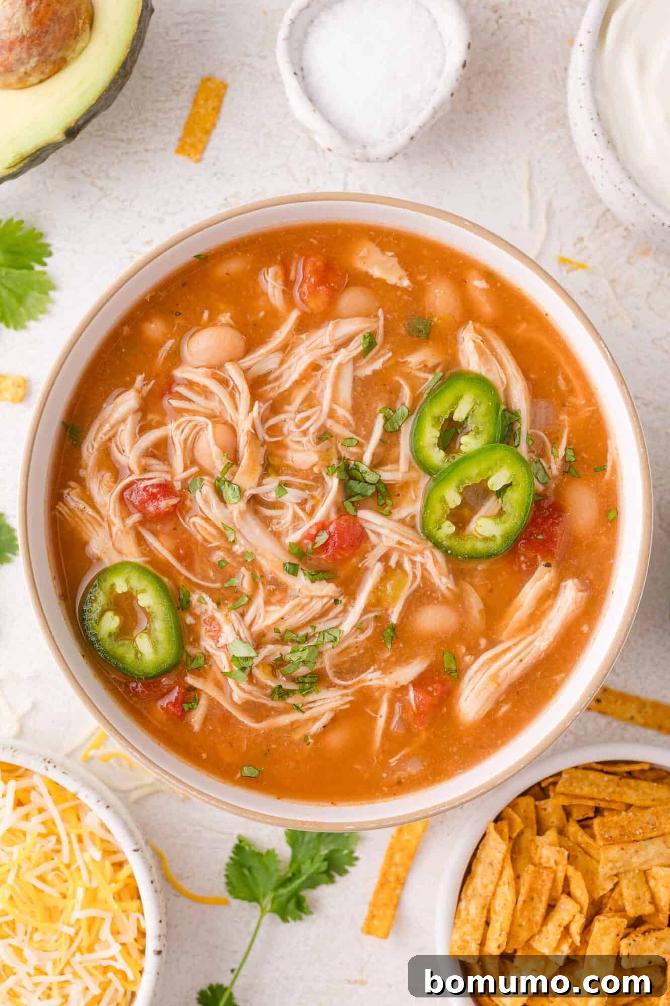 A vibrant bowl of crockpot white chicken chili, garnished with fresh sliced jalapeños and fragrant chopped cilantro. It's elegantly presented alongside small bowls of shredded Monterey Jack cheese, crispy tortilla strips, a perfectly ripe avocado, and a pinch of coarse sea salt, showcasing the chili's rich texture and inviting toppings.
