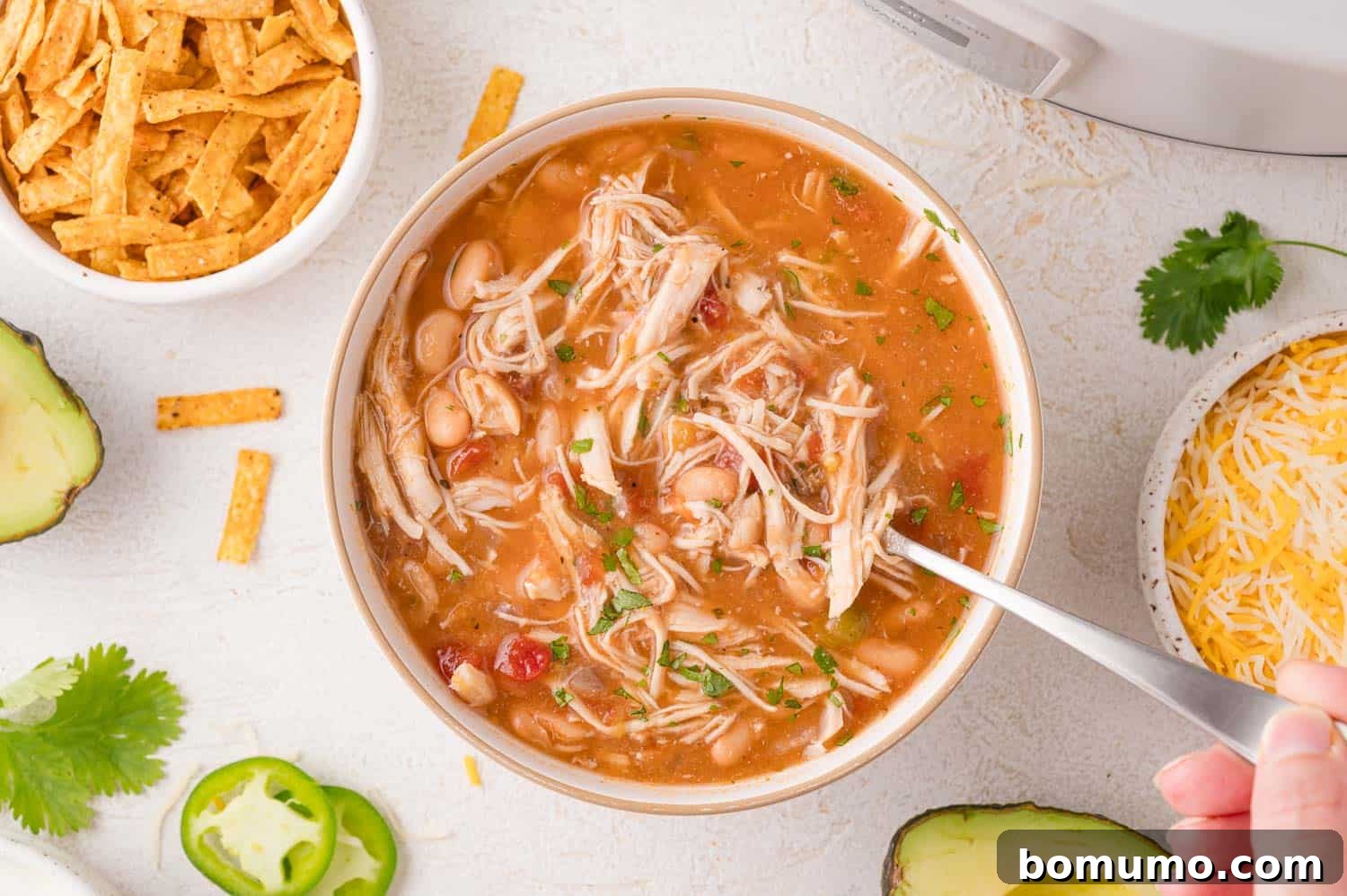 A tantalizing bowl of white chicken chili, rich with shredded chicken, tender white beans, and diced tomatoes, is artfully arranged with an inviting selection of toppings. Surrounding the bowl are small dishes featuring golden tortilla strips, generous piles of shredded cheese, fresh green cilantro, vibrant jalapeño slices, and creamy avocado chunks, all ready to customize your meal.