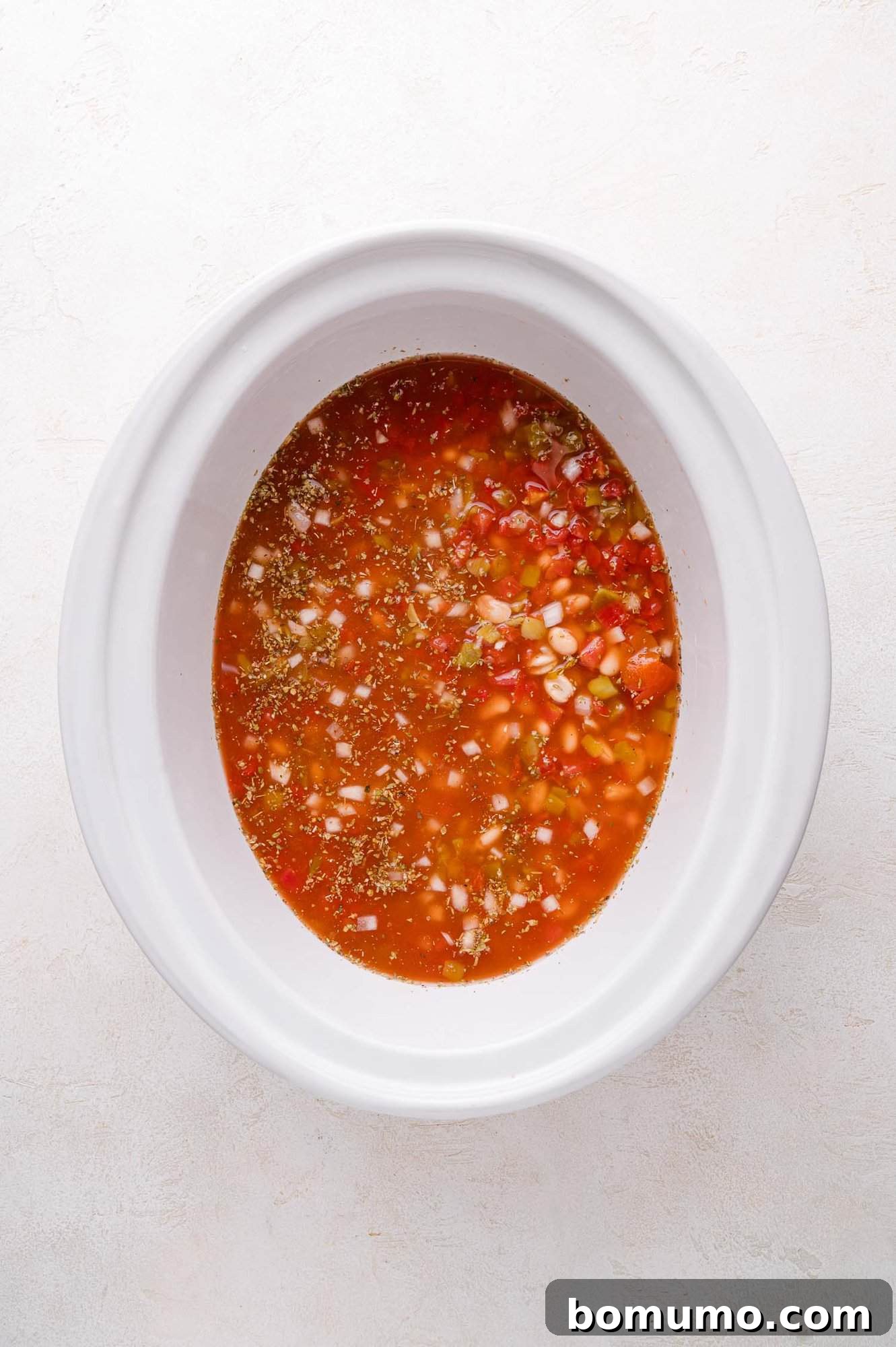 An overhead shot captures a white slow cooker bowl brimming with a vibrant, uncooked mixture for white chicken chili. Visible are diced onions, colorful peppers, and various seasonings, all resting in a rich, tomato-based liquid, ready to begin the slow cooking process.