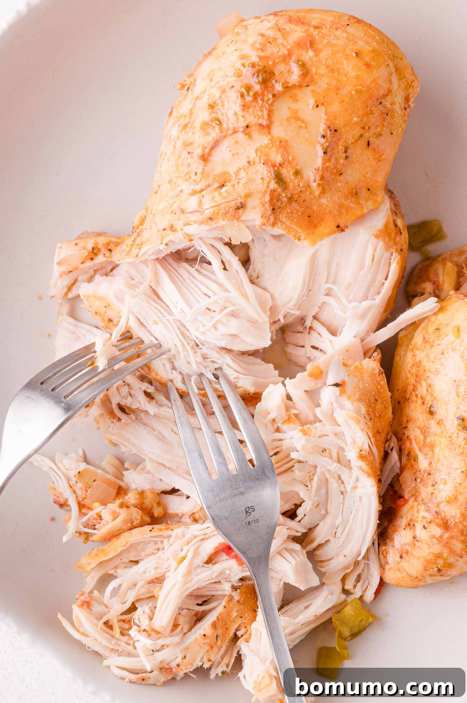 A close-up view of two forks effortlessly pulling apart a perfectly cooked, seasoned chicken breast on a clean white plate. The tender, juicy chicken shreds easily, highlighting its readiness for incorporation into the chili.