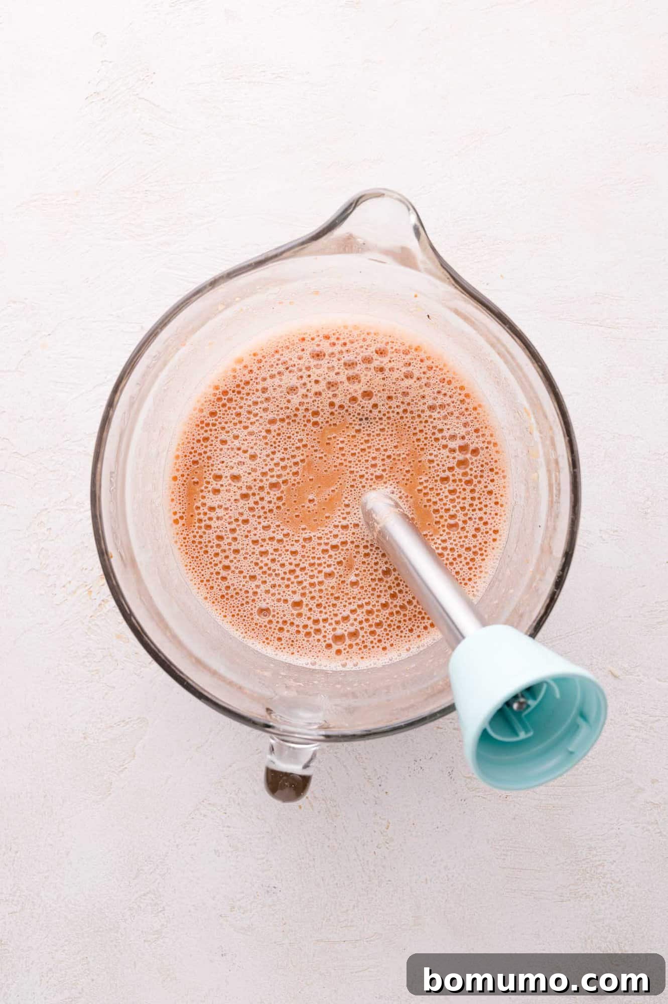 An overhead perspective shows a glass measuring cup containing a frothy, light brown liquid, with an immersion blender fully submerged inside. The scene suggests the process of pureeing a portion of the chili to achieve a thicker, creamier consistency.
