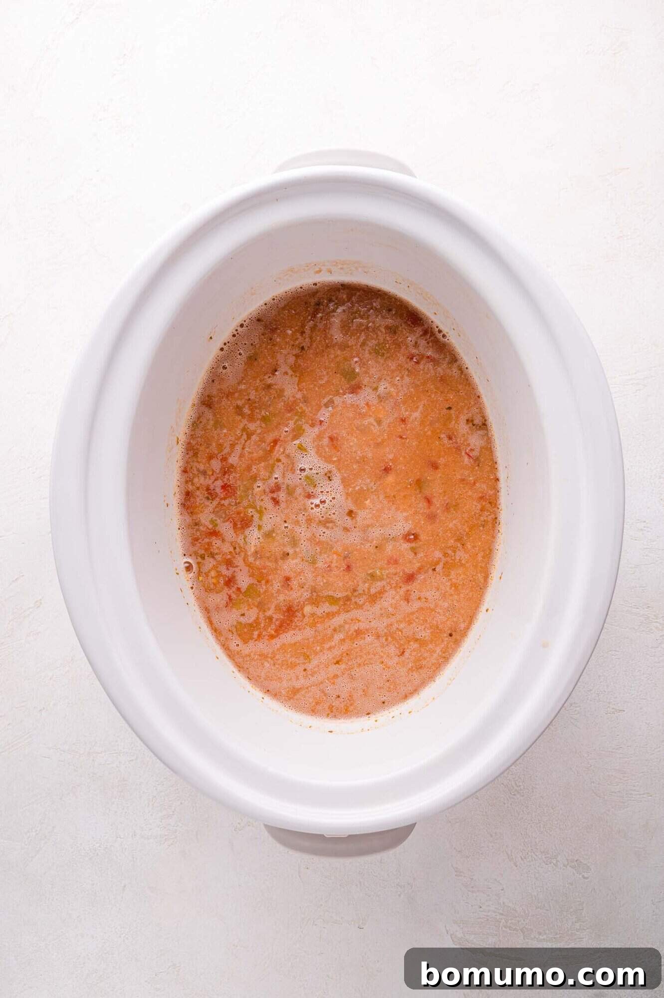 A white slow cooker on a light surface, now containing a smoothly blended mixture of chili ingredients. The consistency appears thicker and creamier, indicating the reincorporation of the pureed portion, creating a rich base for the shredded chicken.