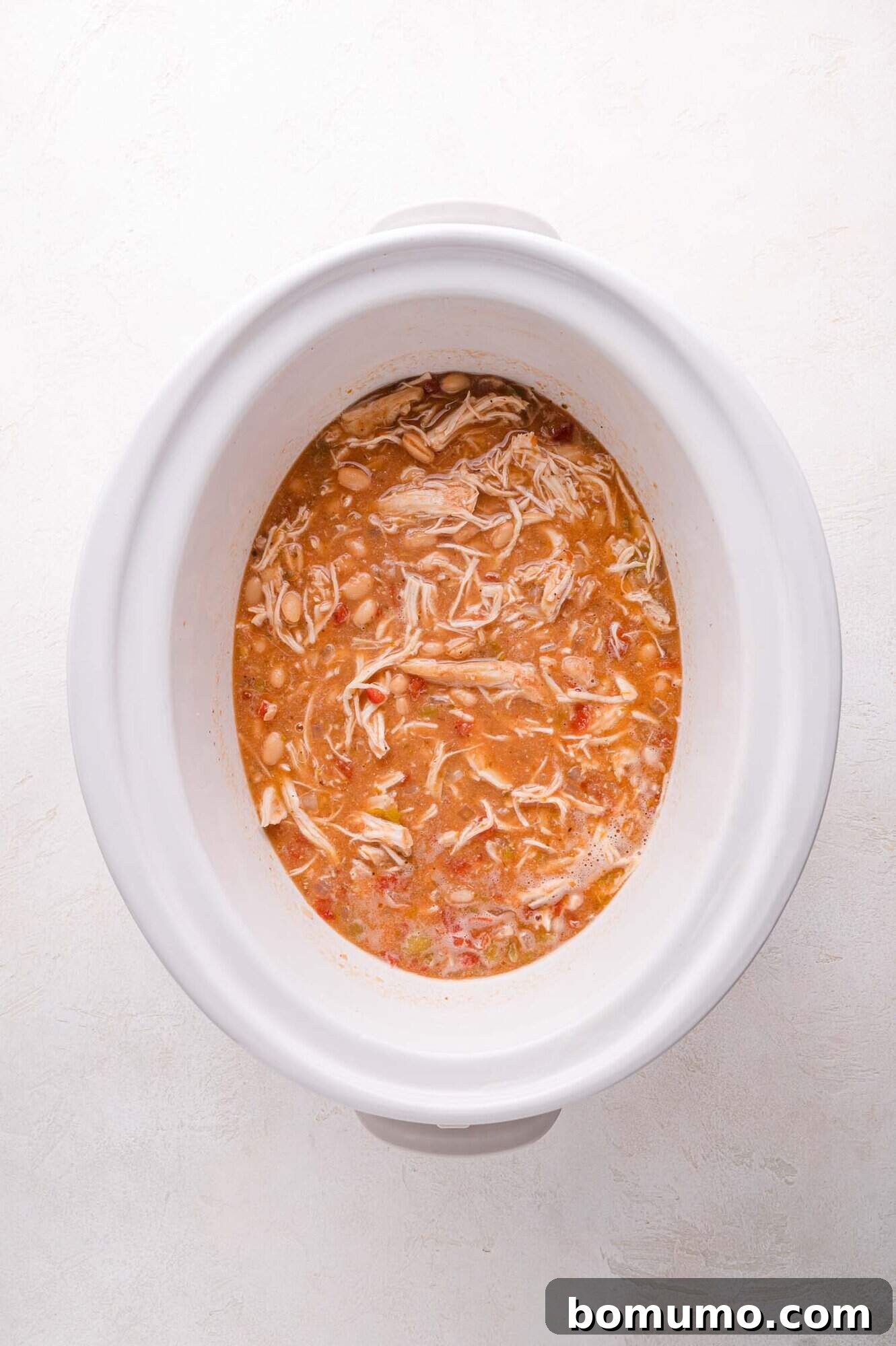 A white slow cooker filled with hearty, shredded chicken chili. The broth is rich, studded with tender white beans and diced tomatoes, and hints of savory seasonings are visible. The shredded chicken is perfectly integrated, promising a satisfying and flavorful meal.