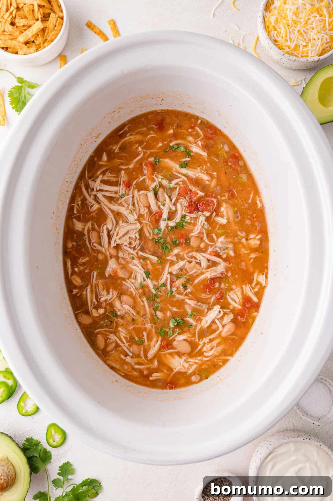 An inviting overhead view of a white slow cooker brimming with finished white chicken chili, surrounded by an appetizing array of topping bowls. Featured garnishes include creamy avocado, generous piles of shredded cheese, a dollop of sour cream, vibrant jalapeño slices, and crispy tortilla strips, all ready for customization.