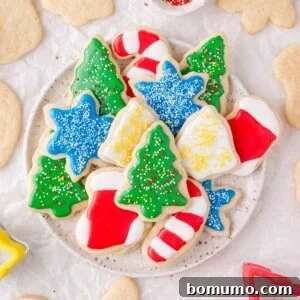 A plate of assorted Christmas cut out sugar cookies decorated with colorful icing in shapes like trees, stockings, candy canes, and stars. Some plain cookies are scattered around the plate.