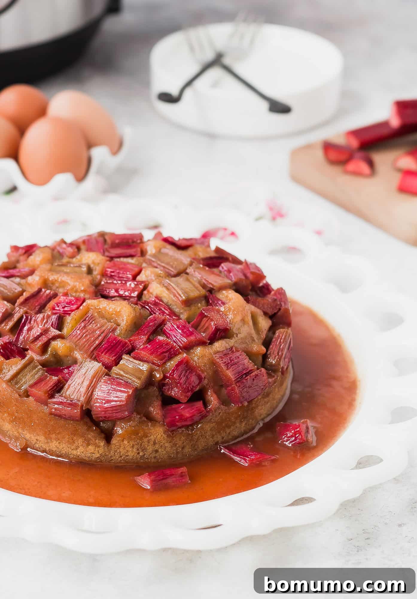 A beautifully presented Rhubarb Instant Pot Cake sits proudly in front of fresh rhubarb stalks, eggs, and the Instant Pot itself, highlighting the fresh ingredients and innovative cooking method.