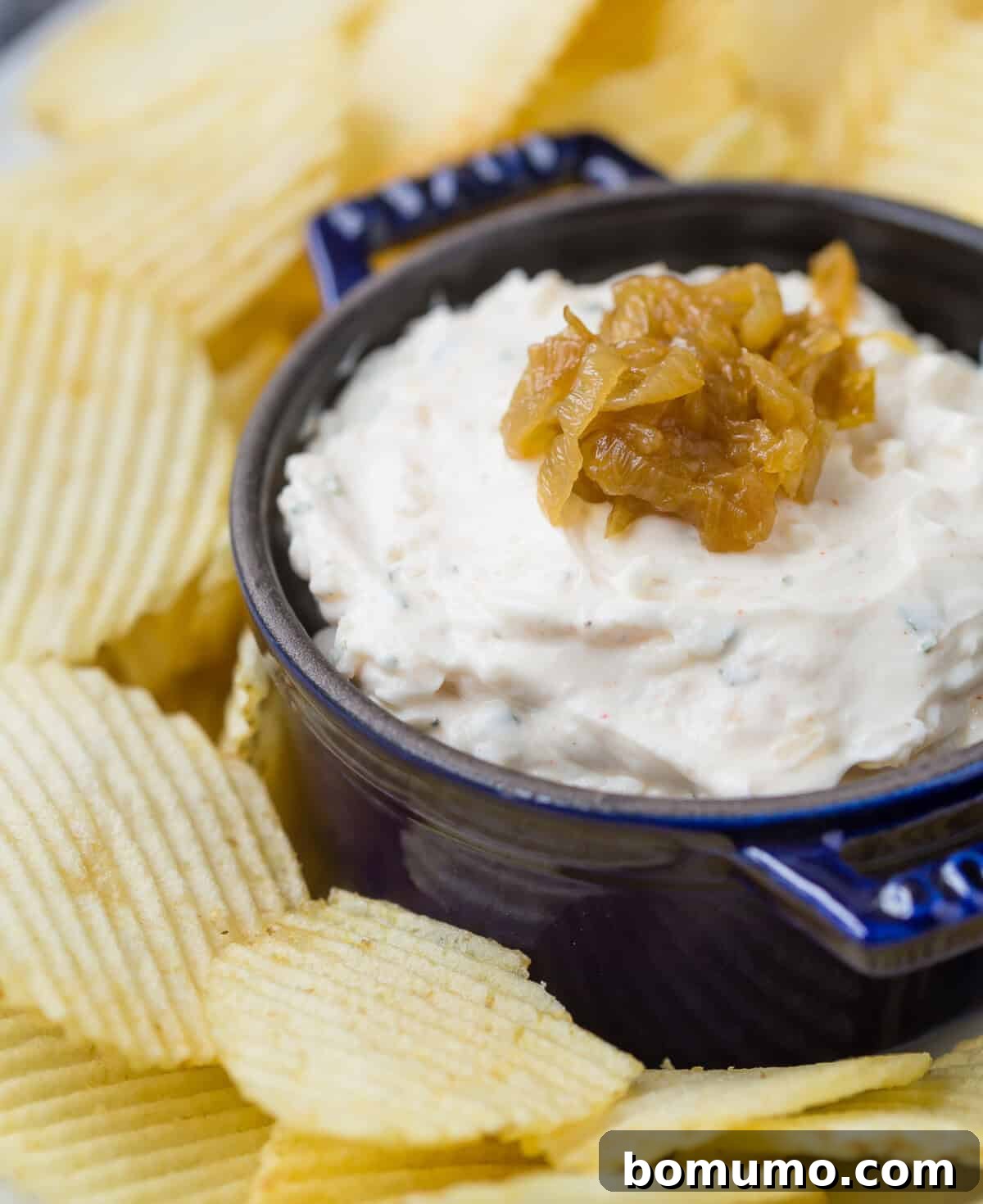 A dark blue bowl of creamy homemade French onion dip topped with a swirl of caramelized onions, surrounded by ridged potato chips.